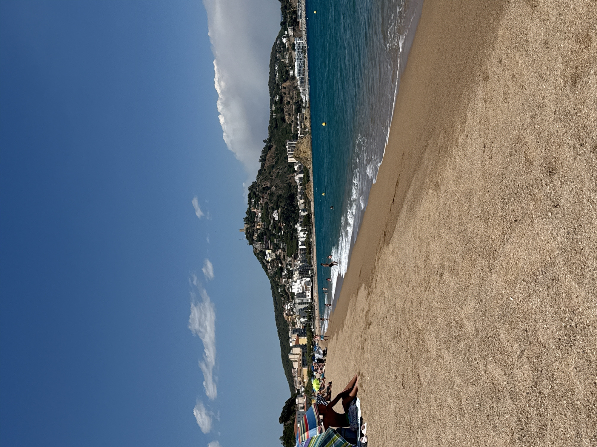 The image captures a sunny day at the beach in Blanes, Spain. The foreground is dominated by a sandy beach with some small pebbles. Several beach umbrellas are set up, and a few people are relaxing or sitting on the sand. In the middle ground, the turquoise water of the Mediterranean Sea meets the shore with gentle waves. Further back, a coastal town sprawls across a hillside, with buildings of various sizes and colors dotting the landscape. Atop the hill, a prominent tower-like structure is visible. The sky is a clear blue with a few scattered white clouds. The scene depicts a typical summer day at a popular seaside resort.