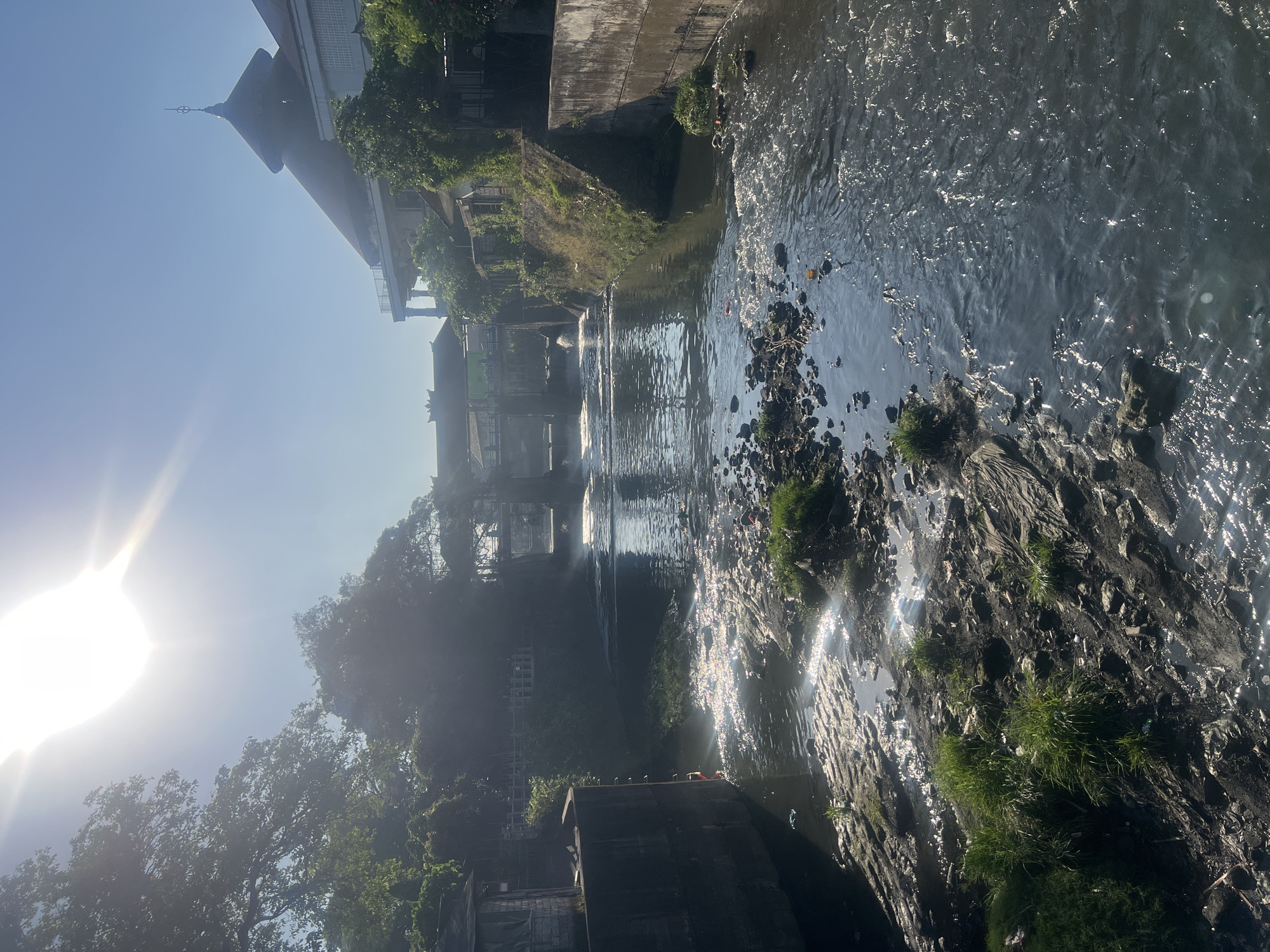 The image shows a canal in Denpasar, Bali, Indonesia, with a building resembling a mosque in the background. The canal is flowing with water that appears to be polluted, with debris and trash visible along the banks and in the water. Lush green trees and vegetation grow along the sides of the canal, with buildings and structures dotting the landscape. The sun is shining brightly, casting a glare on the water and illuminating the scene. The canal appears to be a man-made structure, with concrete walls and a bridge in the distance. The overall impression is of an urban environment that is struggling with water pollution.