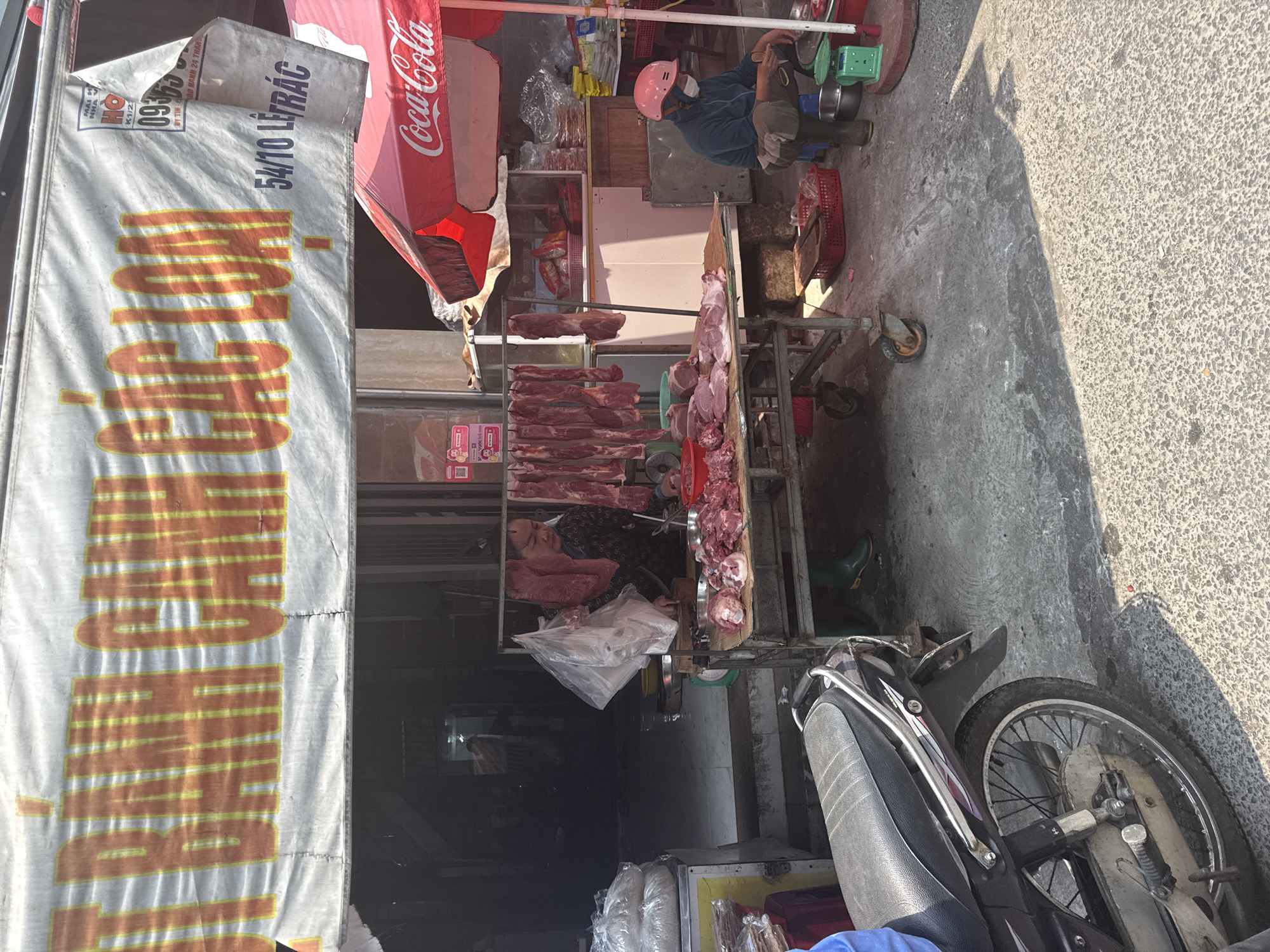 An open-air butcher shop is depicted on a street in Đà Nẵng, Vietnam. A vendor wearing a pink helmet and blue work attire is operating a scale, likely weighing meat for a customer. A table displays a variety of raw meats, with cuts hanging on hooks and arranged on the surface. A woman is standing behind the butcher's table next to the cuts of meat. A Coca-Cola branded canopy is visible above the shop, and a banner with the number "54/10" and the word "LÍTRÁC" is displayed on the side of the shop. A parked motorcycle is in the foreground.