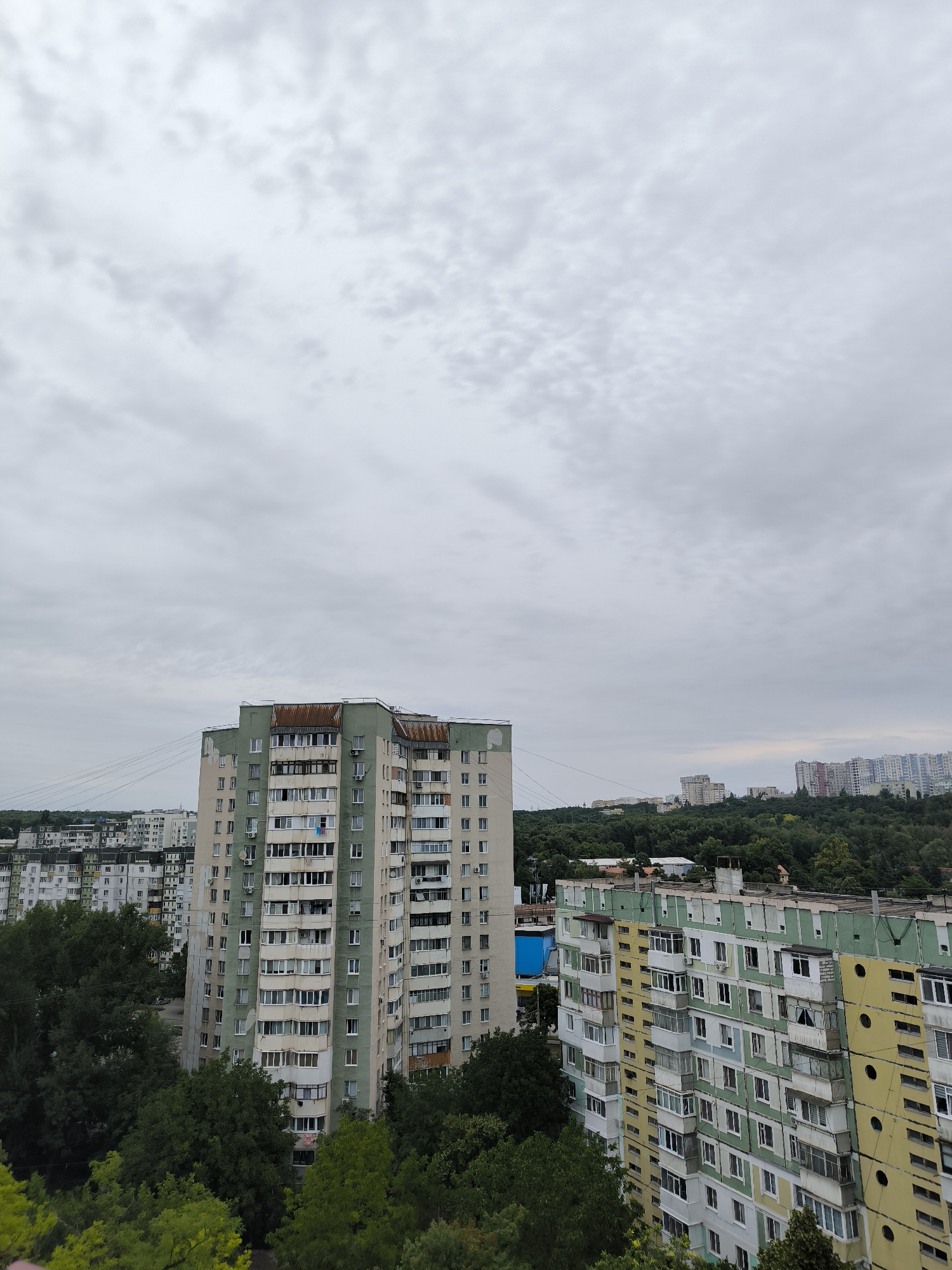 The image shows a cityscape with multiple apartment buildings and trees under a cloudy sky. The foreground features a tall apartment building with a green and off-white facade. Adjacent to it is another multi-story apartment building painted in yellow and green, with numerous balconies. In the background, more apartment buildings and dense green trees are visible. The overall atmosphere suggests an urban residential area in Chișinău, Moldova, possibly a micro-district. The sky is overcast, indicating a potentially gloomy or rainy day. There are no people or discernible actions in the image.