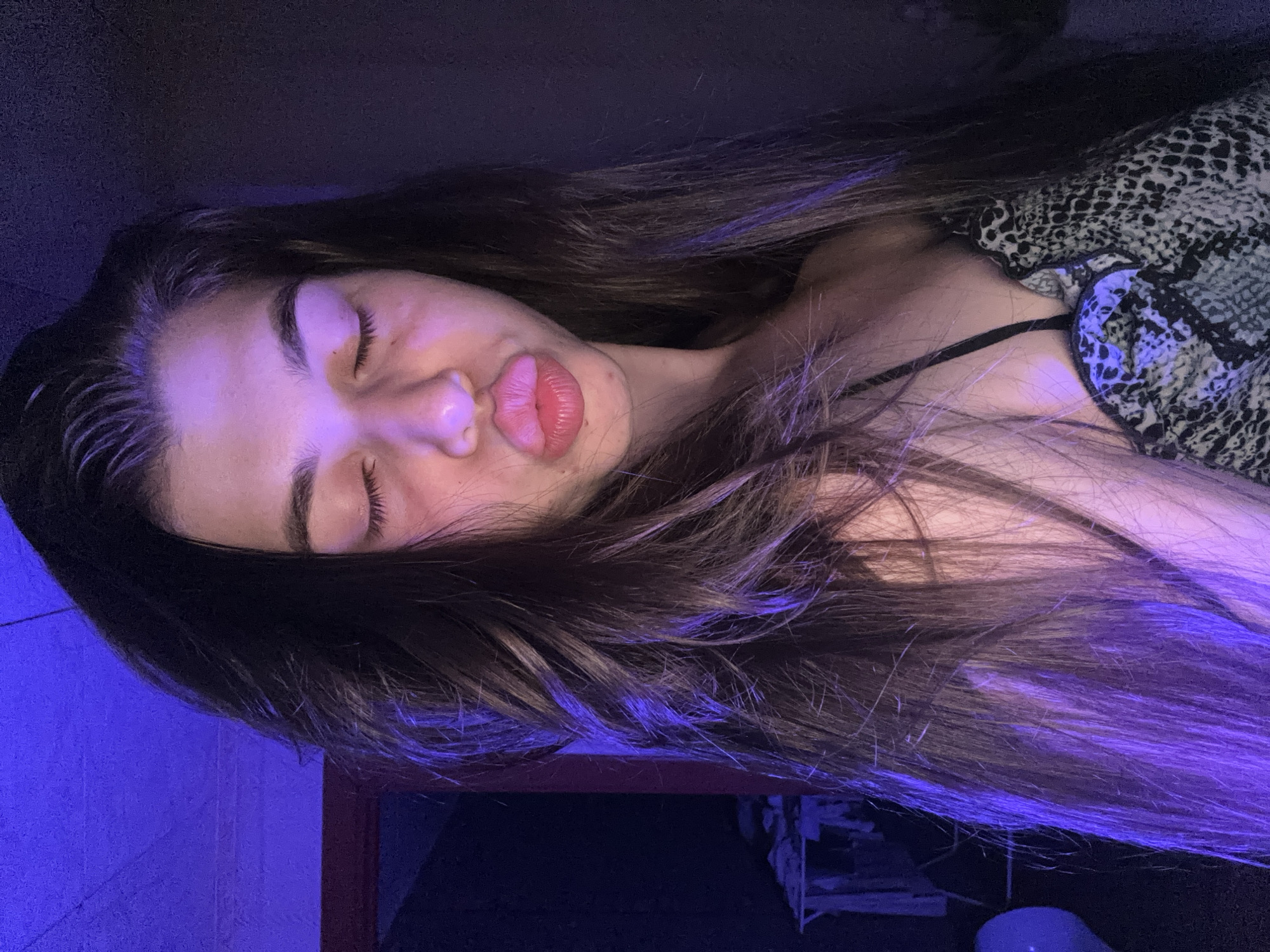 A young woman with long brown hair is lying down, eyes closed, and lips pursed in a pouting expression. She is wearing a patterned top with thin black straps. The lighting appears to be blue and purple, casting shadows on the wall behind her. The location of Мещёры, Russia cannot be verified based on the visual evidence provided.