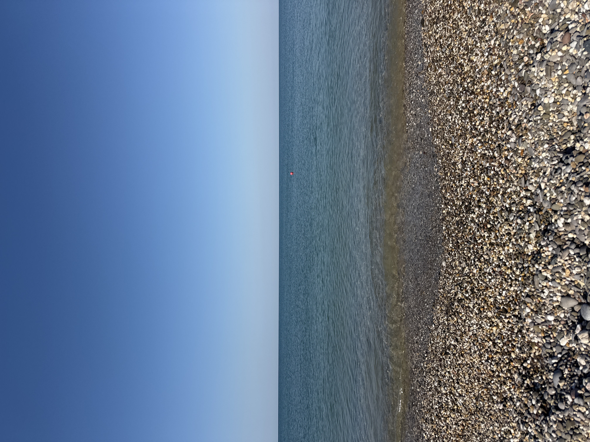 The image depicts a serene coastal scene in Novofedorovka, Ukraine. The foreground is a beach covered in smooth, colorful pebbles, ranging in shades of gray, white, and tan. Gentle waves are lapping at the shore, their clear water revealing the pebbles beneath. In the mid-ground, the vast expanse of the Black Sea stretches out, its surface a tranquil blue under a clear, cloudless sky. A single, small red buoy is visible in the water, marking a point of interest or a designated area. The horizon line is sharp and distinct, separating the deep blue of the sea from the lighter blue of the sky. The overall impression is one of peace and natural beauty.