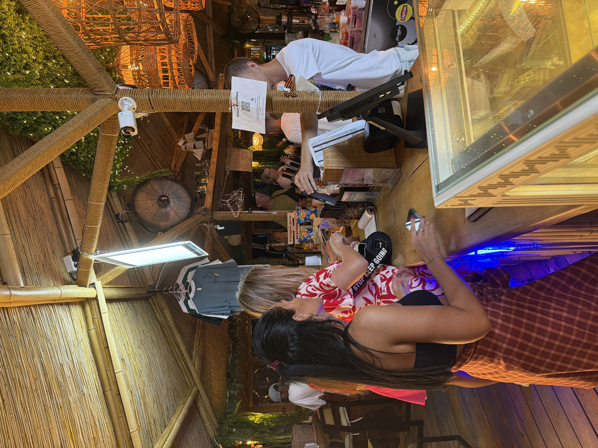 In the city of Sirius, Russia, a busy outdoor bar or restaurant scene unfolds under a thatched roof structure. The setting appears to be a tropical-themed establishment, with bamboo accents and lush greenery decorating the ceiling. A CCTV camera is visible mounted on a rope-wrapped pillar, indicating security measures.
In the foreground, two women are engaged in a transaction at a wooden counter. The woman on the left, with long dark hair pulled back in a ponytail and wearing a black top and a red plaid skirt, is holding a phone. The woman next to her, with blonde hair, is wearing a red and white floral shirt. She appears to be handing over something, possibly payment, to a young man behind the counter. The man, wearing a white t-shirt, is holding a scanner or payment device. A QR code with the text "SOCHI FORNIA" is displayed on a sign attached to the pillar.
Behind the counter, there are various items visible, including bottles of alcohol, snacks, and a cash register. In the background, other patrons can be seen, some standing and others seated. The overall atmosphere suggests a lively and casual social gathering place.