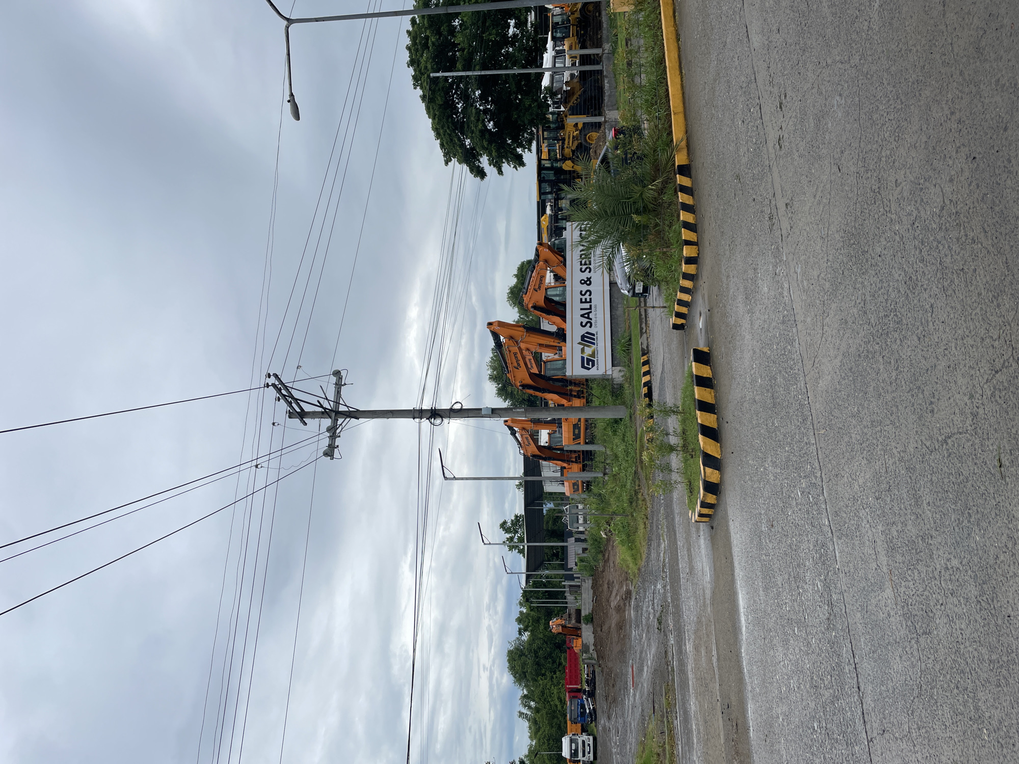 A landscape view of a construction equipment sales and service lot on a cloudy day in Davao City, Philippines. The foreground shows a wet asphalt road with yellow and black striped concrete barriers. In the midground, there are several large orange excavators lined up, with a sign for "GCM SALES & SERVICE". Beyond the excavators, there are more construction vehicles, including trucks and smaller yellow construction equipment, parked under a covered shed. The background features lush green trees and a cloudy sky. The scene appears to be an industrial or commercial area, likely on the outskirts of the city.