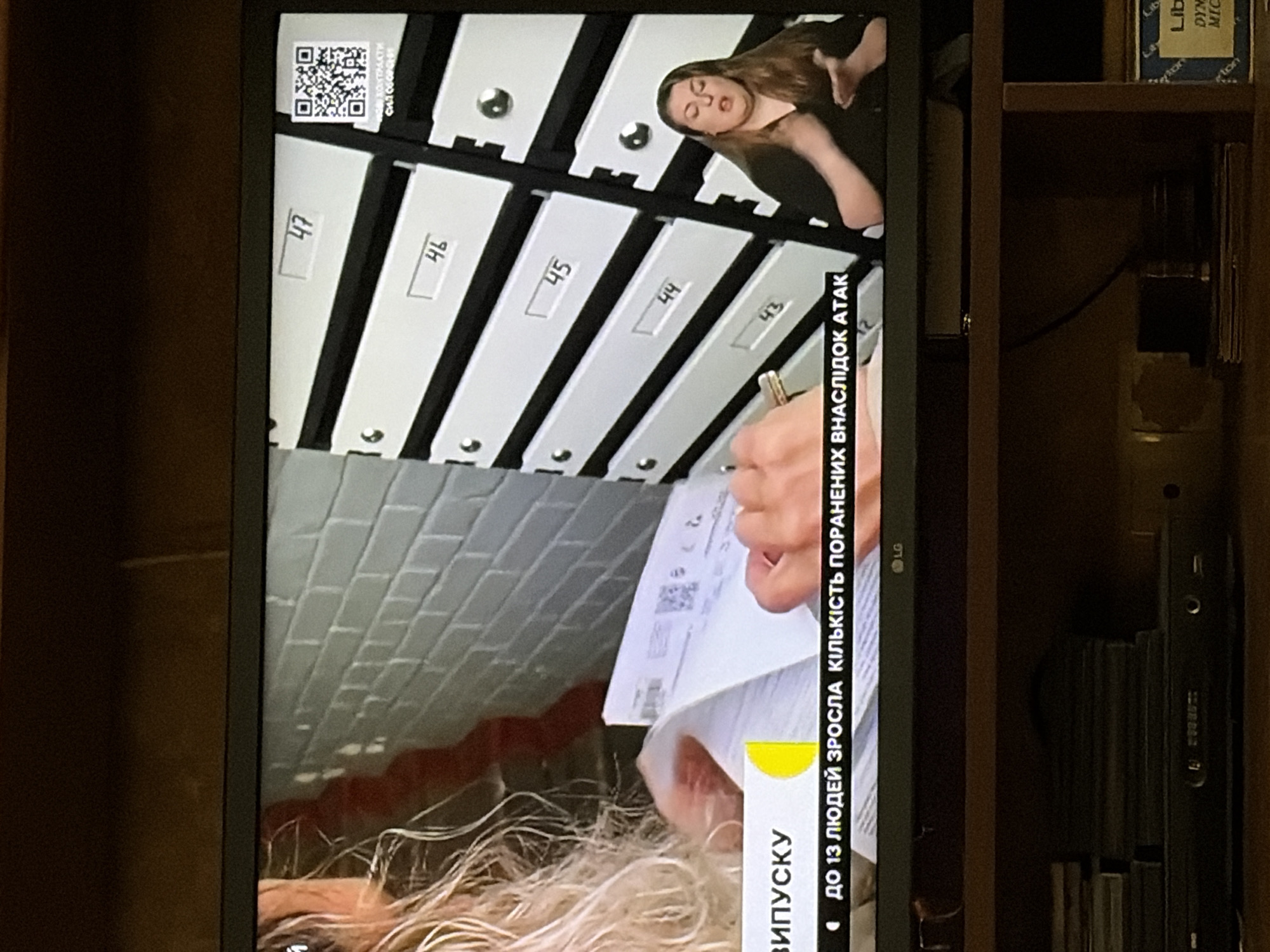The image shows a television screen displaying a news broadcast. On the left side of the screen, a woman with blonde hair is visible, holding a document. To the right, there is a wall with numbered mailboxes, ranging from 43 to 47. A QR code is also visible next to mailbox 47. In the lower part of the screen, a banner reads "ДО 13 ЛЮДЕЙ ЗРОСЛА КІЛЬКІСТЬ ПОРАНЕНИХ ВНАСЛІДОК АТАК," which translates to "Up to 13 people, the number of injured increased as a result of attacks." Below this, the LG logo is visible. On the right side of the screen, a woman is speaking, likely a sign language interpreter. The TV is placed on a shelf with various electronic devices and stacked media below it. The background appears to be a dark wooden shelf.
