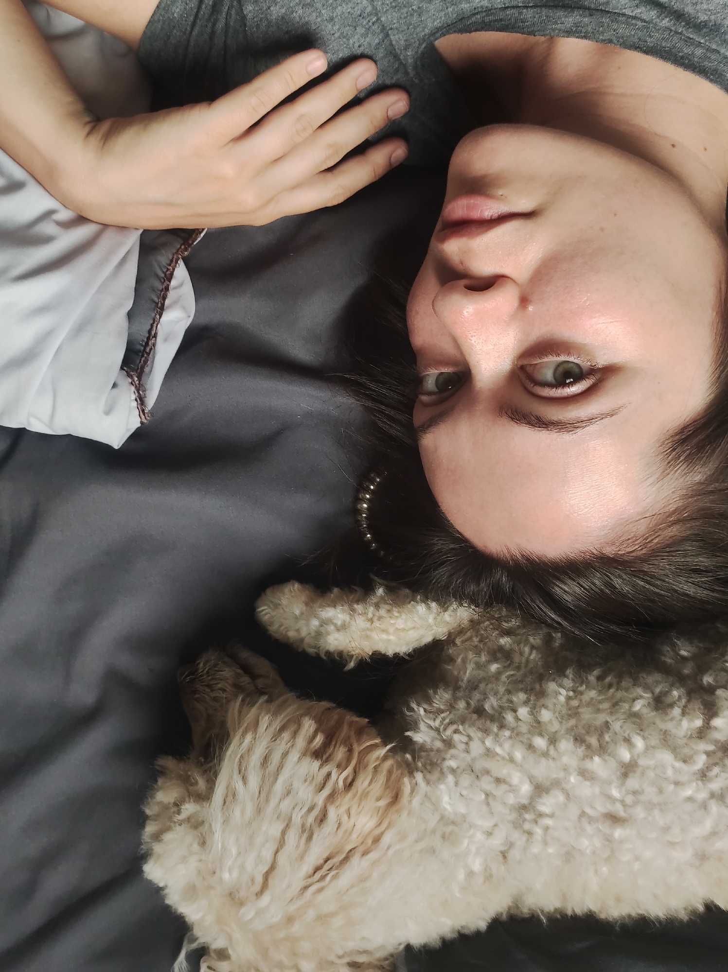 A woman is lying on a grey bed with a small, fluffy, light-brown dog. The woman is fair-skinned with dark hair and green eyes. She is wearing a grey shirt. Her right hand is resting on a white and grey pillow. A pearl necklace is partially visible near her hair. The environment suggests an indoor setting, likely a bedroom. There is no explicit information to suggest a location in the city of Chaikovsky, Russia.