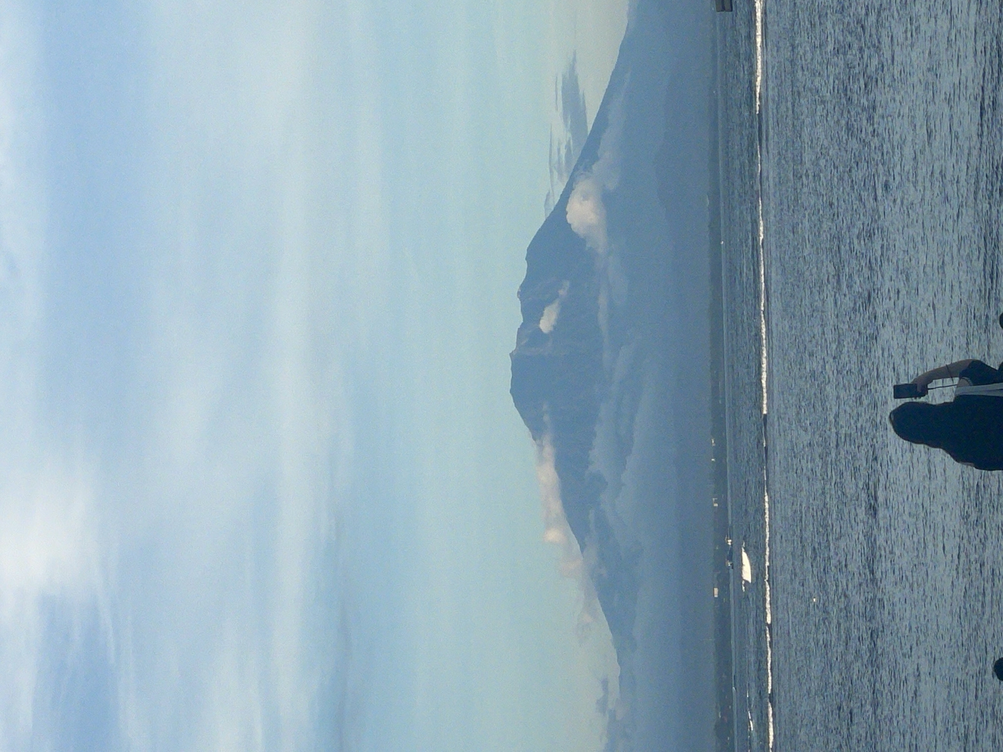 The image, presented sideways and best viewed rotated to landscape orientation, captures a serene outdoor scene featuring a prominent mountain or volcano in the distance. The mountain's dark, imposing silhouette rises under a light blue sky, with some white clouds clinging to its slopes and around its peak, suggesting atmospheric haze or elevation.
In the mid-ground and foreground, a vast body of water, likely a sea or large lake, extends across the width of the frame. The water's surface is textured with gentle ripples and features a striking, elongated horizontal reflection of sunlight, indicating a bright, sunny day. On the far right side of the foreground, a dark, indistinct silhouette of a person is visible, with their arm raised as if holding a phone or camera to capture the scenic view.
The overall setting is tranquil, showcasing natural beauty. The time of day appears to be daytime, possibly morning or late afternoon given the distinct sun reflection on the water. No specific weather conditions beyond partially cloudy are discernible. Given the location context of Denpasar, Indonesia, this view likely depicts one of Bali's significant volcanic mountains from a coastal vantage point. No visible text is present in the image.