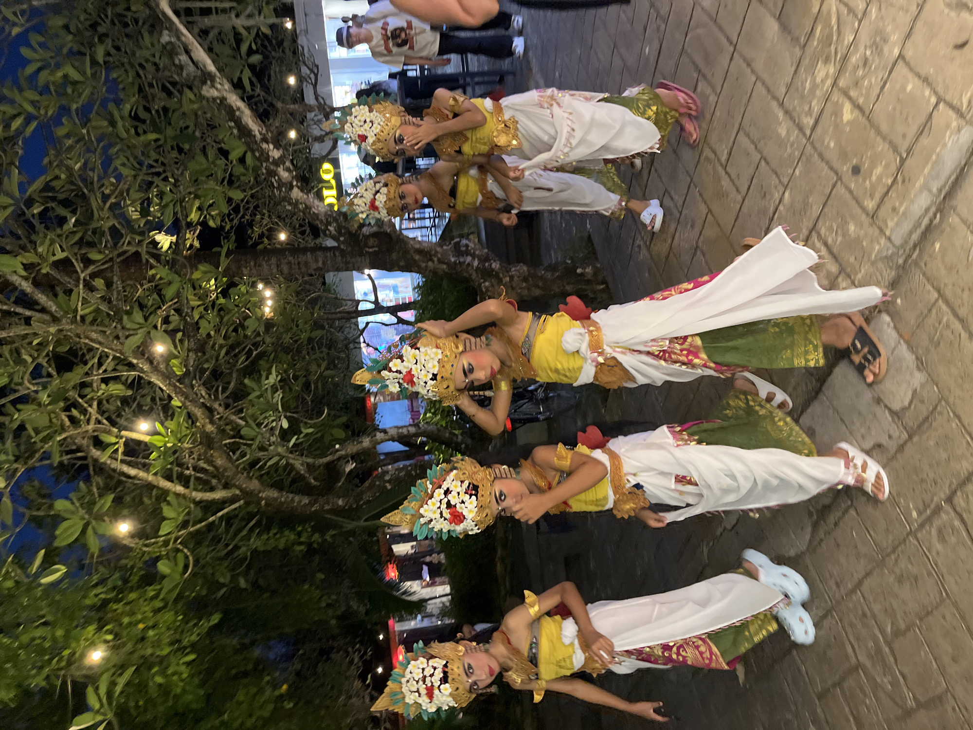 Five young women dressed in traditional Balinese dance costumes stand in a row on a brick-paved area in Kuta Selatan, Indonesia. They wear elaborate golden headdresses adorned with white and red flowers, yellow tops, and long skirts layered with white and green fabric. The dancers pose for the camera, with varying hand gestures and facial expressions. Behind them, a tree with small string lights is visible, and in the background, there are people and buildings. The scene appears to be at night or dusk.