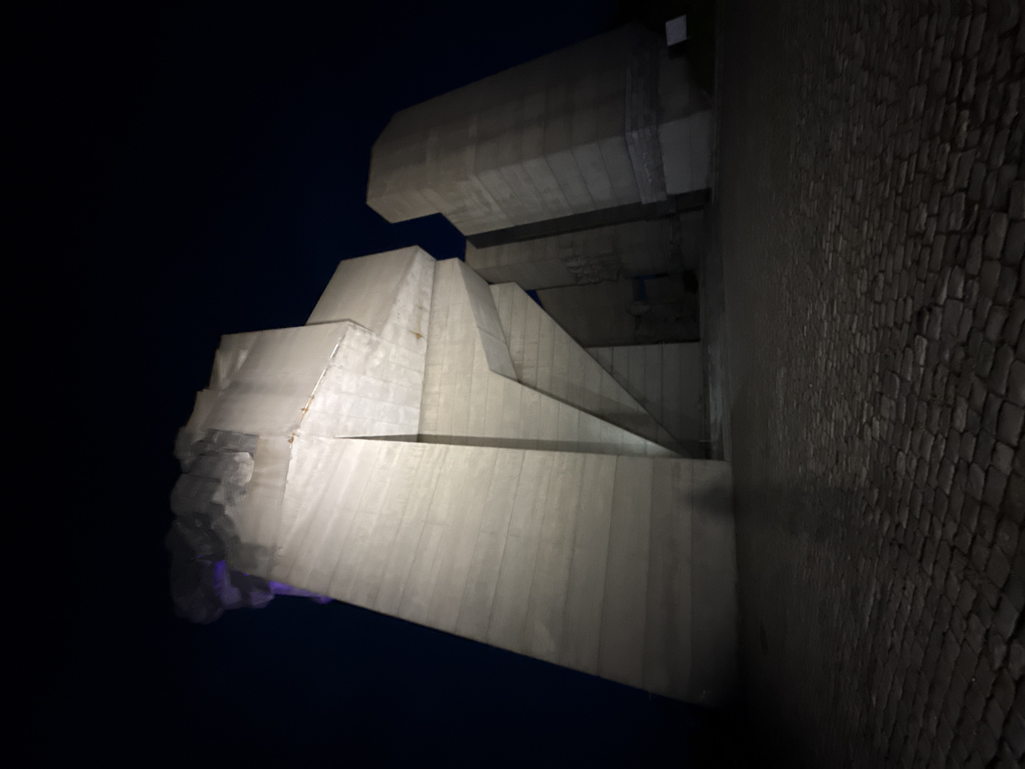 The image shows the Founders of the Bulgarian State monument in Shumen, Bulgaria, at night. The monument is constructed from large, white concrete blocks arranged in a multi-tiered, asymmetrical design. The sky is dark, providing a stark contrast to the illuminated monument. To the right, the ground is paved with dark, textured stones. There are no people visible in the scene. The overall atmosphere is imposing due to the monument's scale and stark lighting.