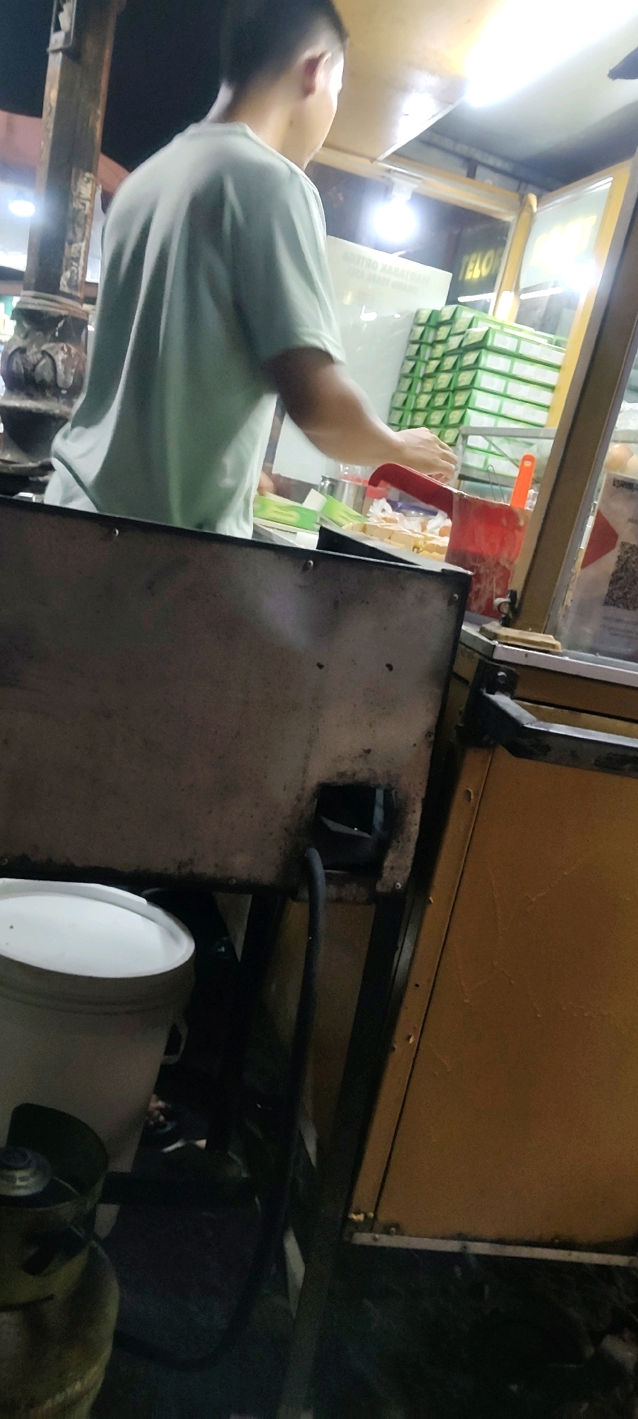 The image shows a street food vendor in Cibatu, Indonesia, preparing food. A person wearing a light green t-shirt is standing behind a food cart, with their back to the camera. They appear to be in the process of serving food, as their hands are reaching towards a container of food. The food cart is equipped with a grill, a container of food, and a stack of green boxes. In the foreground, there is a white bucket and a gas cylinder. The scene is dimly lit, suggesting that it is nighttime or early morning. The background is blurred, but it appears to be a street scene with lights.