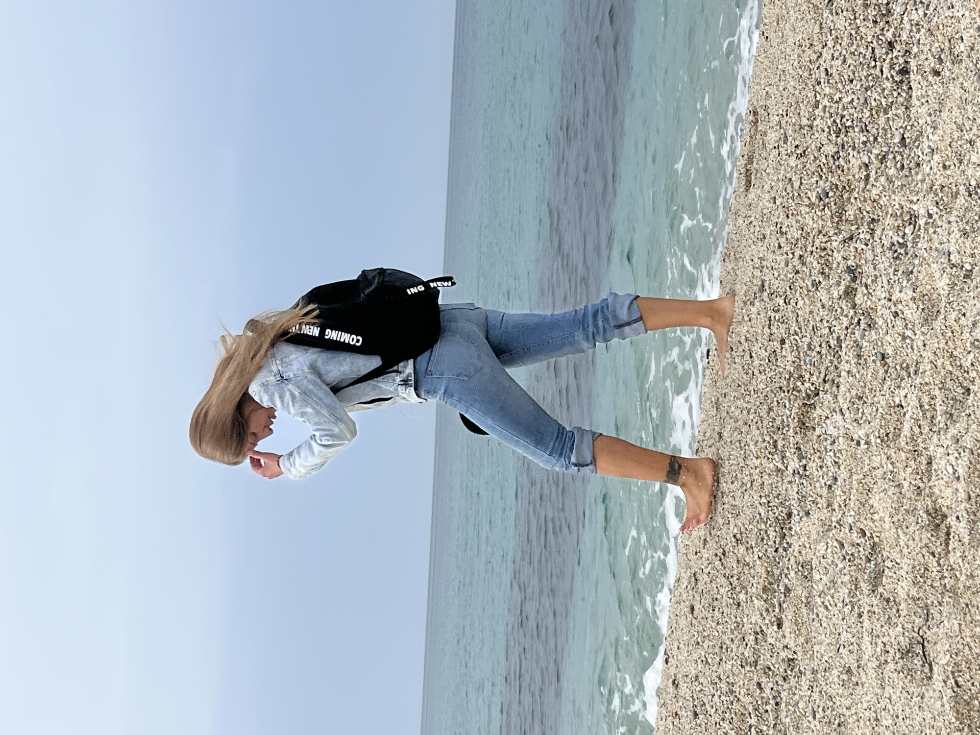 The image shows a person standing barefoot on a pebbly beach next to the sea, under a clear sky. The person, appearing to be a young woman with long blonde hair, is dressed in light blue denim jeans, rolled up at the ankles, and a light-colored denim jacket. She has a black backpack on her back. Her posture suggests she is looking downwards, possibly at the sand or water, with her hands near her face.
The scene features a wide expanse of light blue sky, a calm greenish-blue sea with small waves gently breaking on the shore, and a beach composed of small, light-colored pebbles and shells. The horizon line is visible, separating the sea from the sky. The lighting is bright and even, indicating daytime, likely morning or early afternoon, with no visible clouds.
A notable detail is the white text "COMING NEW" visible on the black backpack. One of the person's ankles appears to have a small, dark tattoo. The location context provided is Alanya, Turkey, which aligns with the coastal setting. The overall atmosphere is peaceful and natural, capturing a moment of contemplation or engagement with the coastal environment.