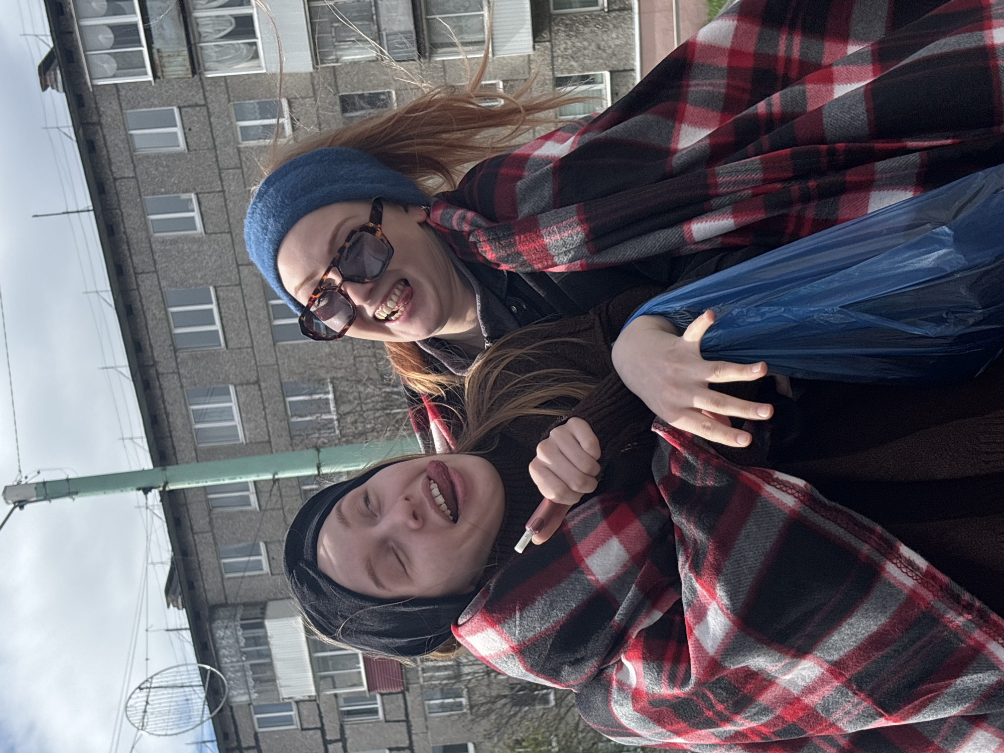 Two young women are huddled together, appearing to be enjoying themselves outdoors. One woman has red hair and is wearing a blue beanie and sunglasses. She has her mouth open, seemingly laughing or smiling. The other woman has dark hair and is wearing a black beanie; she is sticking her tongue out. Both women are wrapped in a red and black plaid blanket. The woman with red hair is holding a crumpled blue plastic bag in her hand. The background features a gray apartment building with multiple windows and an antenna on the roof, suggesting this is in a residential area. The scene takes place in Kachkanar, Russia.