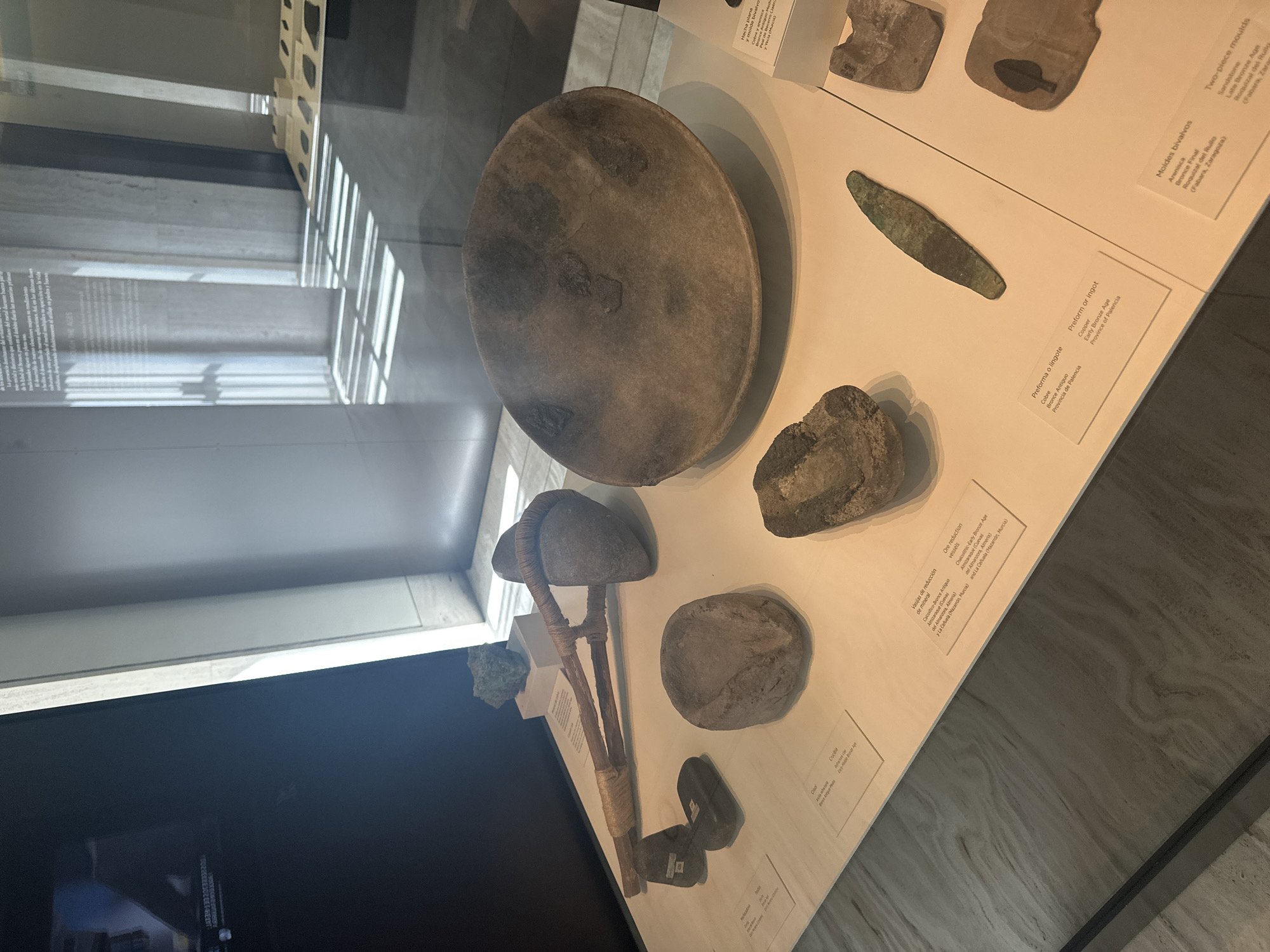 This image displays an exhibit of ancient artifacts, likely from a museum, presented on a white surface behind a glass barrier. The artifacts include stone tools, a large ceramic bowl, and what appears to be a metal ingot or preform, along with some stone molds. The background shows a modern architectural interior with concrete pillars and angled sunlight casting striped shadows. The text labels suggest the location is in Madrid, Spain, specifically within the Museo Arqueológico Nacional (National Archaeological Museum), as indicated by the Spanish text on the labels and the museum's known collection. The artifacts are arranged in a curated manner for public viewing, with informational cards placed beside them, describing their material, age, and origin. There are no people visible in the frame, and no actions or interactions are taking place. The lighting is a mix of ambient interior light and strong directional sunlight.