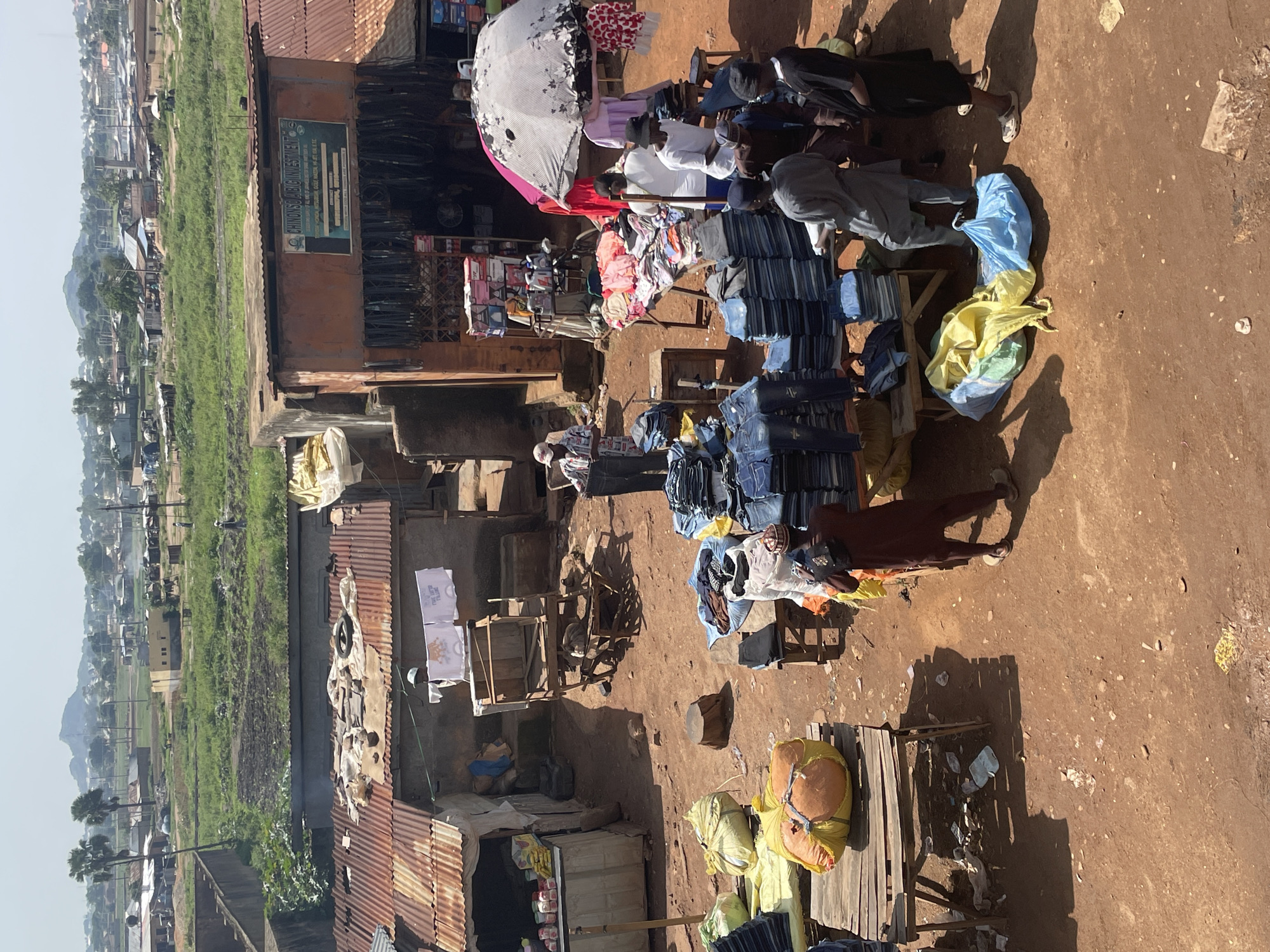 An outdoor market scene is depicted, with multiple stalls and people engaged in commerce. In the foreground, several wooden tables are laden with merchandise, primarily stacks of blue jeans and folded clothing. Large, colorful sacks, one yellow and orange, another blue and yellow, are placed on the ground near the tables. A man in a maroon tunic and a patterned hat is standing with his back to the camera, holding a mobile phone. To his right, several men in similar attire are gathered around a table displaying folded clothes. A large, partially collapsed umbrella, white with black floral patterns, is set up over a stall where clothing is displayed.
Behind these foreground stalls, there are additional structures with corrugated metal roofs, suggesting more vendors. One building displays a sign with text in English. Further in the background, the market gives way to a more rural or undeveloped area with green fields and scattered buildings, leading to a distant landscape with a prominent hill or mountain. The ground is predominantly reddish-brown earth, with scattered debris. The sky is bright and appears to be daytime, with some clouds visible. The overall impression is a bustling, informal marketplace.