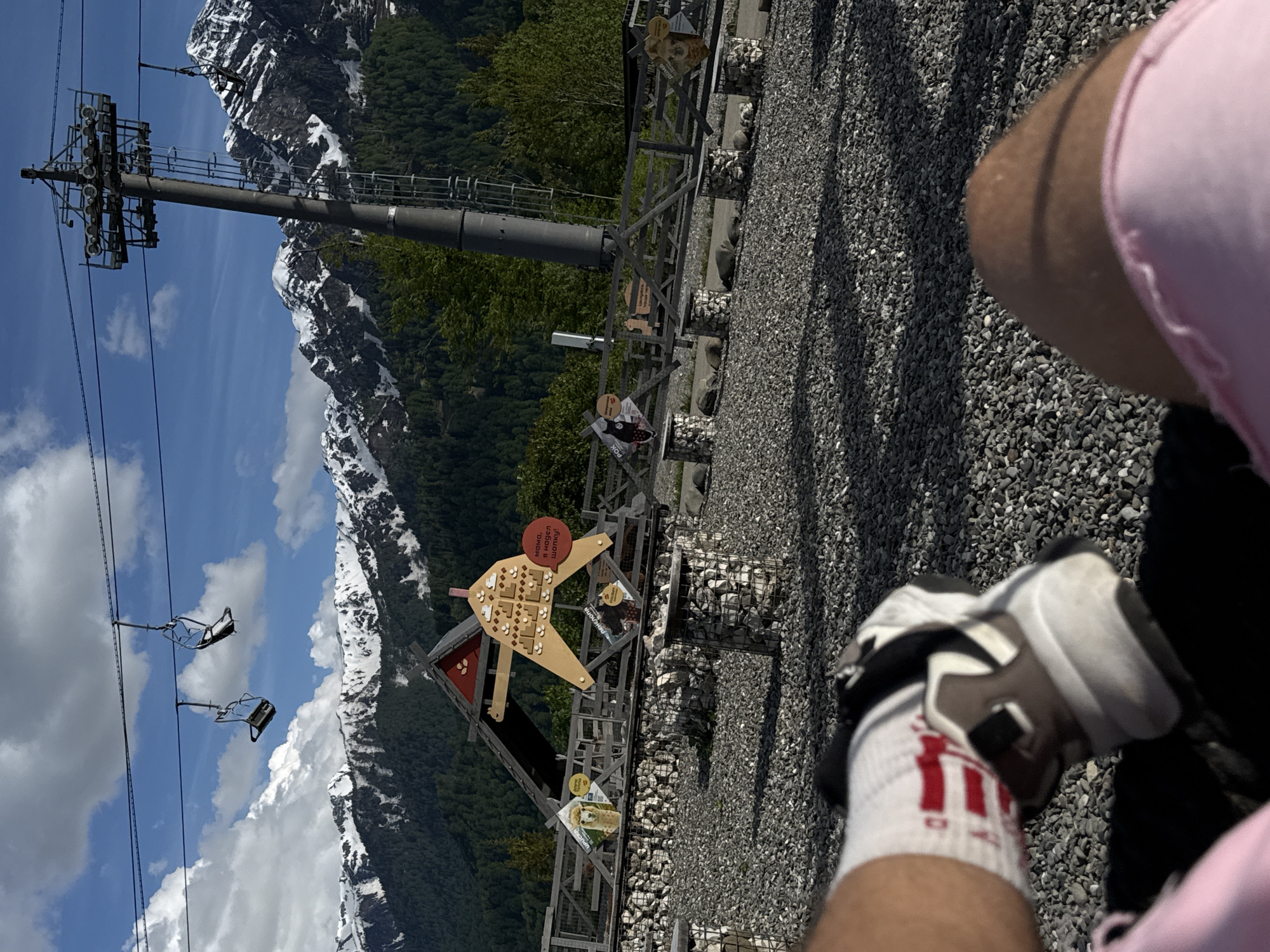 The image, oriented vertically, captures a bright daytime scene in a mountainous recreational area. In the immediate foreground, the lower body of a person is visible, showing thighs clad in pink shorts, white athletic socks with red stripes, and white, grey, and black sneakers. The ground beneath is covered in small, grey gravel.
The midground features a wooden play structure or interactive exhibit, possibly for children. It's a large, light-brown cutout, somewhat abstract, with smaller wooden pieces affixed to it. A red circular sign on this structure displays the Russian text: "Мама, я надел шапку!" (Mom, I put on a hat!). Below, several smaller signs are visible, one depicting a marmot-like animal and partially showing the Russian text "Ледниковый период" (Ice Age). Another sign clearly shows "АЛЬПИНА" (ALPINA). The area around the exhibit is fenced with wood and includes gabion-style rock cages.
In the background, a tall, grey ski lift pylon stands prominently on the left, supporting cables from which several empty chairlifts hang. Beyond the pylon and the green tree line of a dense forest, majestic snow-capped mountains dominate the horizon under a clear blue sky with scattered white clouds. The bright sunlight casts distinct shadows, indicating a clear and pleasant day. The presence of ski lifts and snow-capped peaks, combined with the recreational exhibits, suggests a mountain resort or park setting in Russia, designed for outdoor leisure and activities. The overall impression is one of a serene and enjoyable natural environment with man-made attractions.