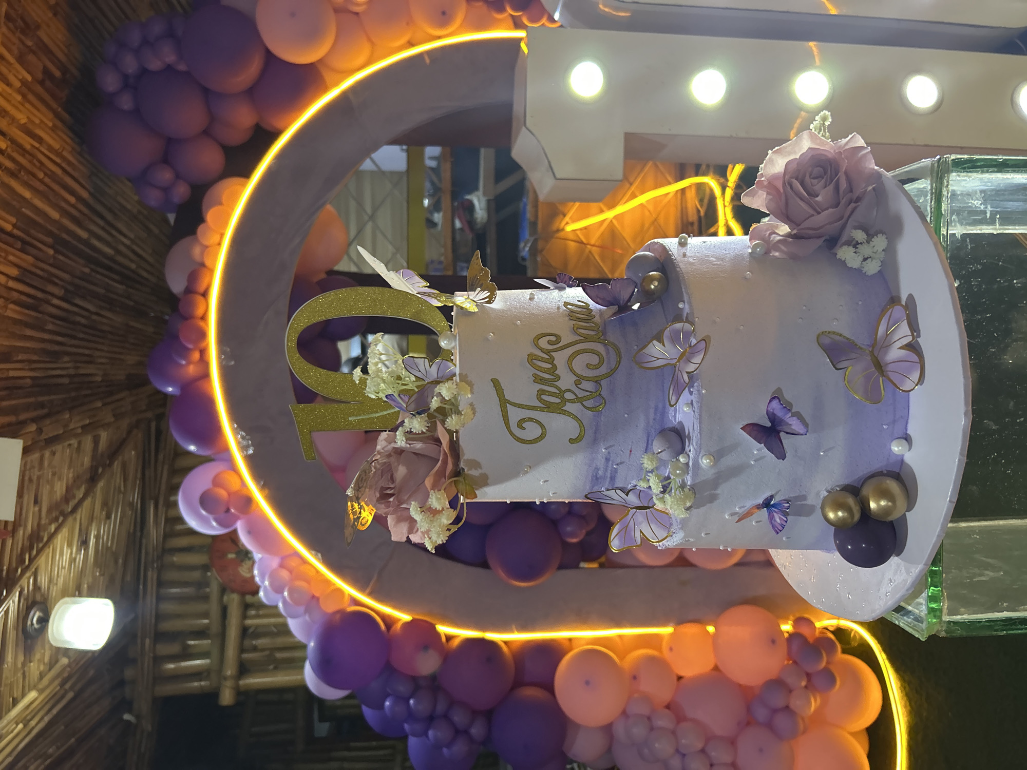 A two-tiered cake with purple and white frosting is adorned with edible butterflies, pearls, and a pink rose. The cake is decorated with the names "Tara & Sara" in gold lettering. A large gold numeral "10" sits atop the cake. The cake is placed on a clear stand with a green base, and is part of a party setup that includes a balloon arch made of purple and pink balloons, and a large illuminated numeral "1". The backdrop consists of bamboo structures and a thatched ceiling. The image does not provide any information or context about the city of Bar Jirgi Summa, Nigeria.