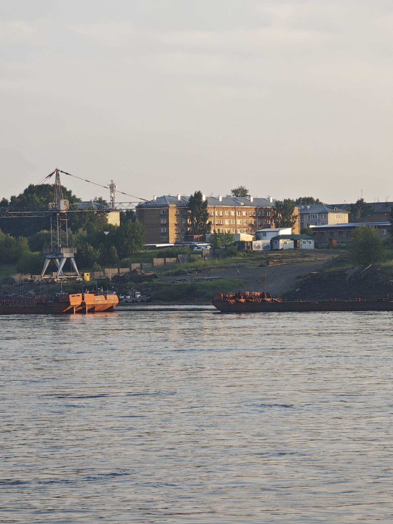 The image depicts a scene along the Angara River in Lesosibirsk, Russia. In the foreground, the calm river water reflects the pale, overcast sky. Two orange barges, weathered and appearing to be under repair or docked for a long period, are prominent on the water's surface. One barge is closer to the viewer, showing its stern with two large, rounded pontoons. The other barge is further away, appearing as a longer, flatter vessel.
The background features a raised riverbank with a mix of industrial and residential elements. A large, industrial crane with a distinctive X-shaped base stands tall on the left side, indicating a port or shipbuilding facility. Behind the crane and along the embankment, several multi-story brick apartment buildings are visible, characteristic of Soviet-era architecture. Smaller structures, including what appear to be sheds or outbuildings, are scattered amongst the buildings. Dense green trees and vegetation line the riverbank and surround the structures. A chain-link fence runs along parts of the embankment. The overall impression is one of a working river town, with signs of both industrial activity and everyday life. There are no visible people in the image.