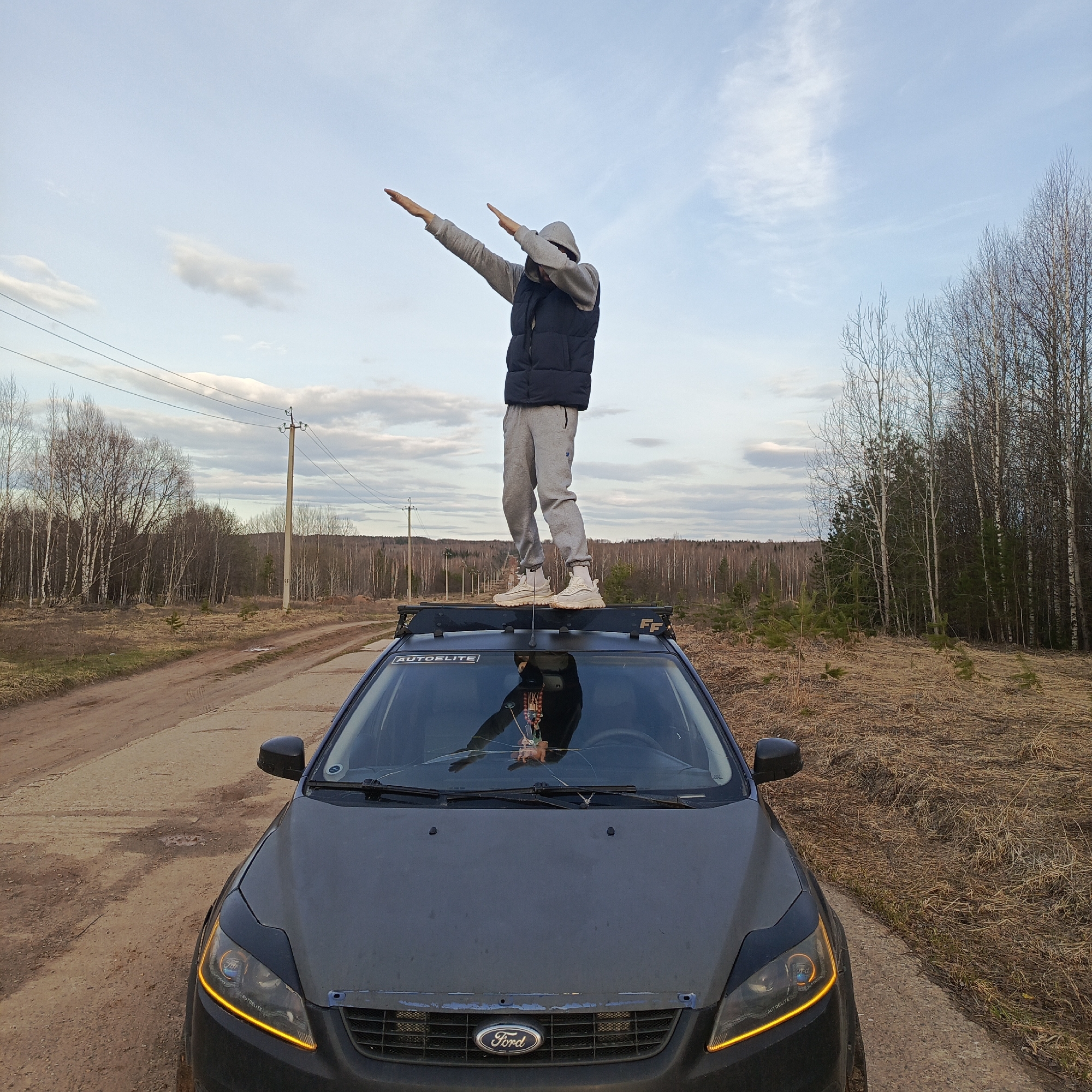 The image shows a man standing on the roof of a dark-colored Ford Focus car, dabbing with both arms extended and head lowered. He is wearing a grey hooded sweatshirt, a dark vest, and grey sweatpants. The car has a roof rack. The windshield of the car is cracked. The background shows a dirt road and a landscape with trees, including bare trees, suggesting a location in the late fall or early spring. Power lines are visible above. Based on the surroundings, the scene appears to be located in a rural or undeveloped area. The car is parked on a dirt road, with fields and trees on either side. The image was most likely taken in the city of Марковский, Russia based on the prompt.