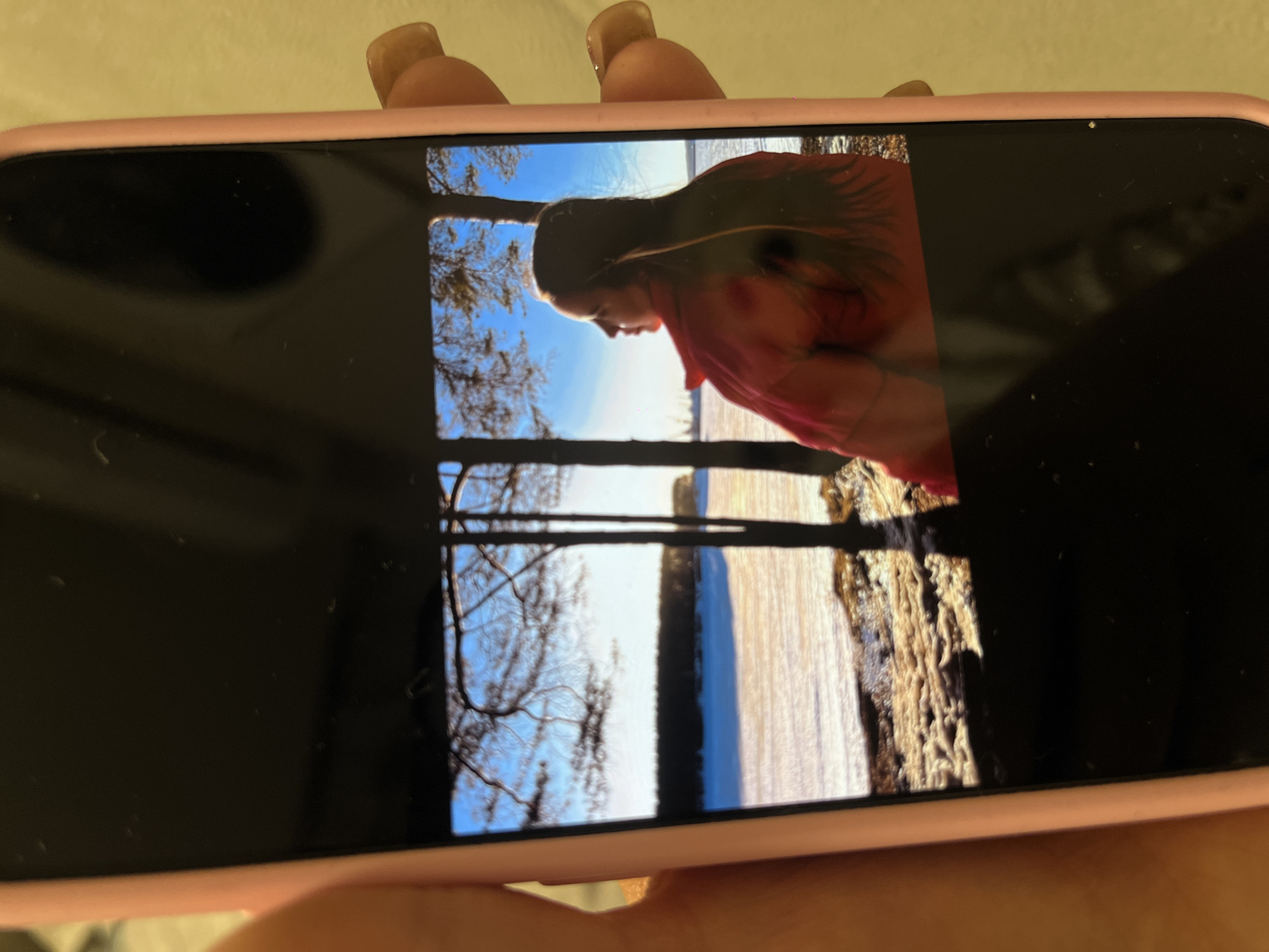 The media file captures a close-up view of a smartphone held in two hands, against a light-colored background. The phone is encased in a soft pink cover, and parts of the fingers or fingernails of the person holding it are visible at the top and bottom edges of the frame.
The smartphone screen displays a vivid outdoor photograph, oriented sideways (90 degrees clockwise from vertical). The central subject of this displayed image is a young woman, shown in profile, looking towards the left side of the frame. She has long, dark hair, partially illuminated by the light, and is wearing a reddish-orange or pink long-sleeved top or jacket. Her expression appears calm or contemplative.
The background of the displayed photograph features a bright, clear blue sky dominating the upper portion. Below the sky, a wide, light-colored expanse stretches out, which could be a frozen body of water or a snow-covered landscape, reflecting the bright daylight. Silhouetted dark, bare tree branches are visible on the left side of the image, extending upwards. Another vertical dark element, likely a tree trunk, stands towards the center-right. The strong lighting suggests a sunny daytime, possibly in winter or early spring, given the bare trees and potentially frozen landscape. The overall mood conveyed by the image on the screen is serene.
Notable details include several small dust particles or smudges visible on the black areas of the phone screen. There is no visible text within the displayed image or on the phone's interface. The natural elements, such as bare trees and open, possibly frozen expanses, are consistent with landscapes found in regions like Krasnodar, Russia, particularly during colder months.