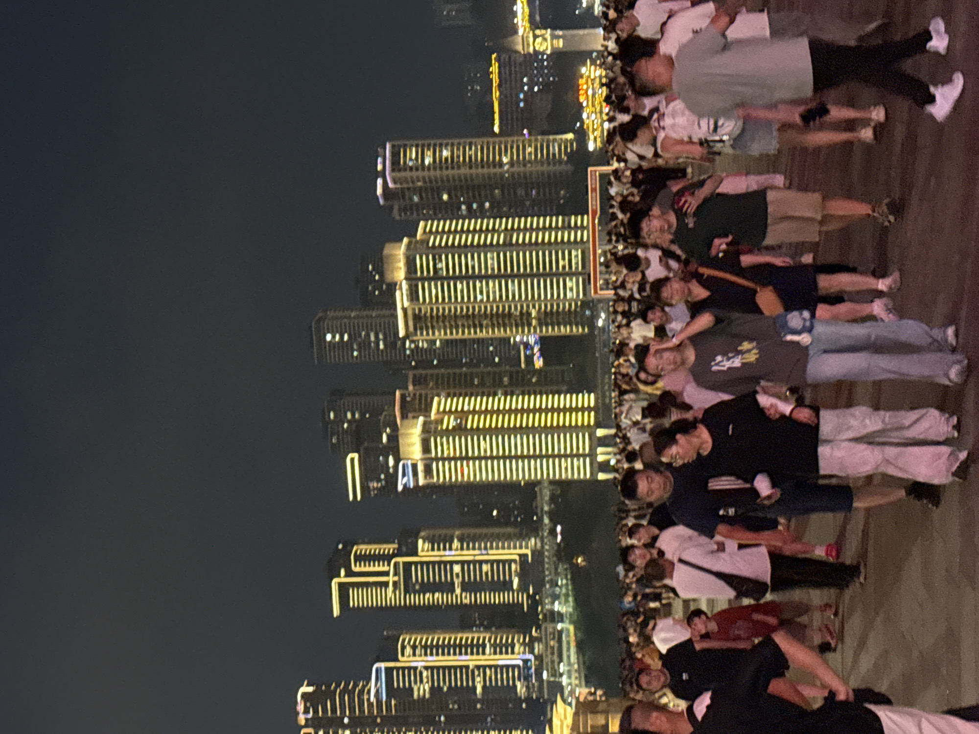 This image captures a nighttime scene in the Yuzhong District of China, characterized by a densely populated urban environment illuminated by artificial lights. A large crowd of people is seen walking on a paved area, suggesting a public gathering or event. In the background, numerous tall, modern buildings are brightly lit with yellow and white lights, forming a prominent cityscape. The architecture of the buildings appears to be a mix of contemporary high-rises. The atmosphere is festive and active, with many individuals dressed in casual attire. The lighting creates a stark contrast between the illuminated buildings and the darker sky, indicating it is nighttime.