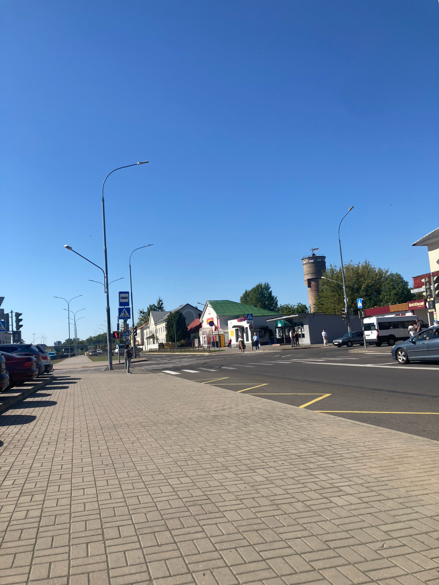The image captures a street scene in Brest, Belarus, on a clear, sunny day. The foreground is dominated by a beige brick-paved sidewalk. To the left, parked cars are visible, casting shadows on the sidewalk. The street itself is paved with asphalt and features a crosswalk with white stripes. Several streetlights with modern designs are spaced along the sidewalk.
In the middle ground, a woman wearing a patterned shirt and light-colored pants stands on the sidewalk, facing the street. She appears to be waiting or observing. A bus stop sign and a pedestrian crossing sign are visible nearby, mounted on a pole. Across the street, a row of buildings lines the road. These buildings vary in style and color, with some having green roofs. A distinctive tall, round brick water tower with a cap is visible in the distance, set against a backdrop of trees. Several vehicles, including cars and a white van, are present on the street. The sky is a clear, vibrant blue.
