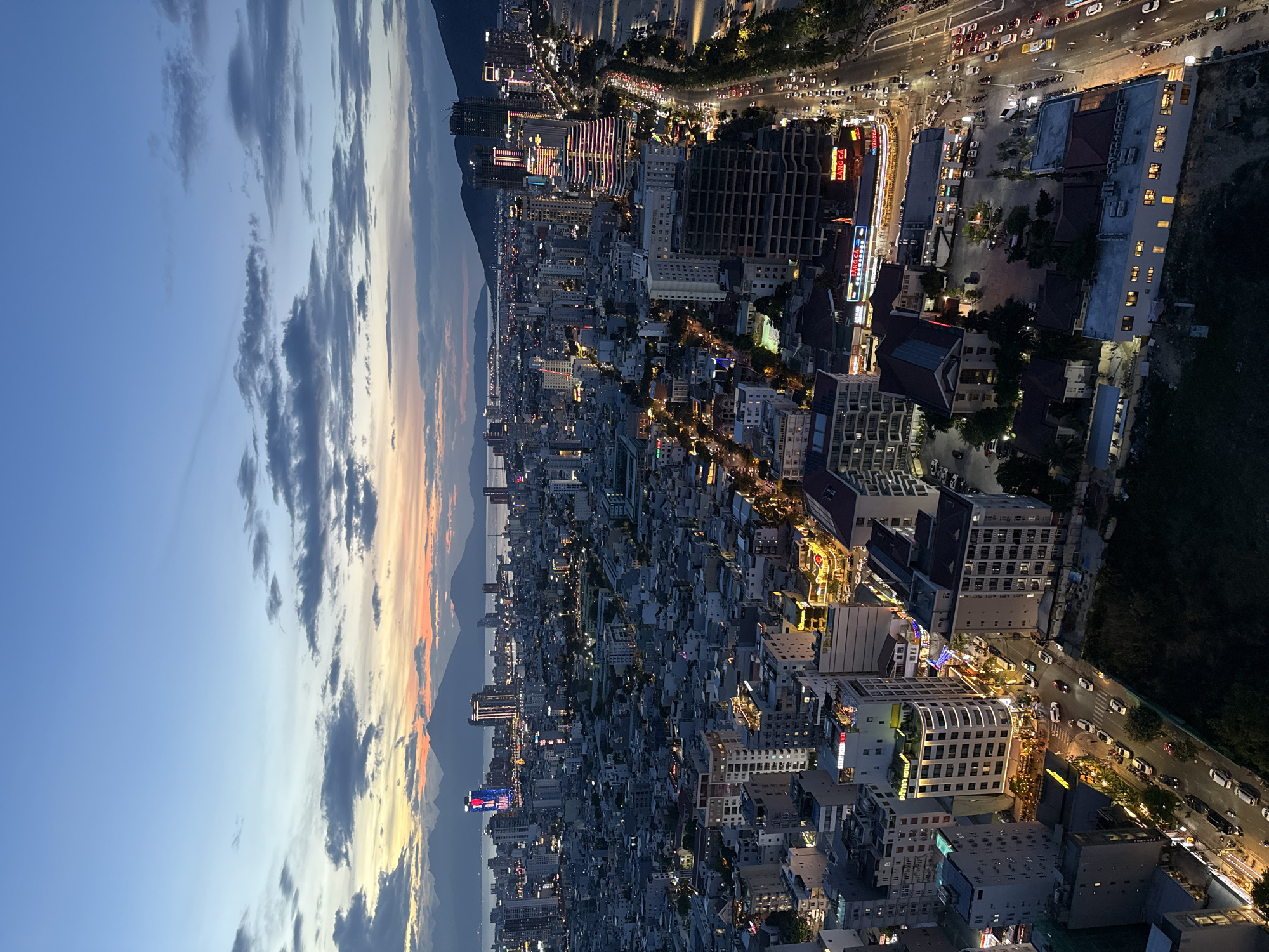 This media file presents a high-angle, wide shot of a sprawling city at dusk, transitioning into night, viewed from a tall building or elevated position. The image is oriented vertically, showing the horizon line across the middle.
The upper portion of the original image (left side in the current orientation) depicts the evening sky, a gradient of deep blue fading into warm oranges, yellows, and purples near the horizon where the sun has set. Scattered clouds, predominantly dark gray and white, streak across the sky, catching the last light of the day.
Below the horizon (right side in the current orientation) lies a dense urban landscape, stretching towards distant mountains and a body of water, possibly the ocean or a large river, where a bridge can be faintly discerned. The city is illuminated by countless artificial lights from buildings, streets, and vehicles, creating a vibrant, intricate pattern against the deepening twilight.
Numerous buildings of varying heights and architectural styles populate the cityscape, some glowing with interior lights, others highlighted by exterior signage. A prominent multi-lane road snakes through the city, bustling with vehicles whose headlights and taillights form bright streaks. Along this road and within the urban blocks, smaller buildings, trees, and other urban infrastructure are visible.
Visible text includes "LANG CA" on multiple prominent signs, possibly indicating restaurants or businesses, one appearing as "LANG CA SEAFOOD". The overall impression is one of a lively, modern city continuing its activities as night falls. Given the context, this is a nocturnal view of Đà Nẵng, Vietnam.