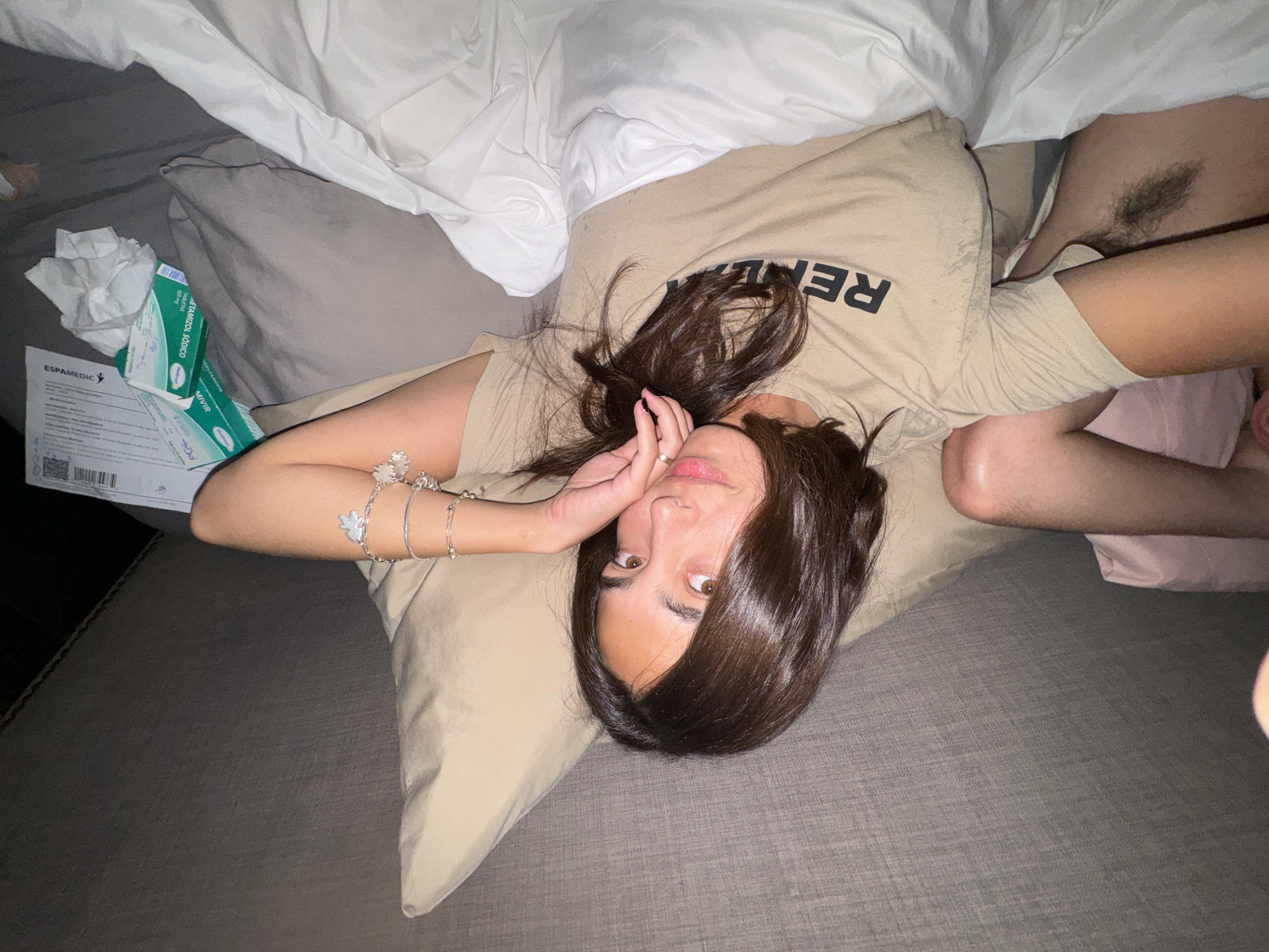 A woman is lying on a gray bed. She is resting her head on a tan pillow. She is wearing a tan t-shirt with black lettering. Her right arm is outstretched, and she is wearing multiple bracelets. Her left hand is touching her face. White bedding is visible in the upper portion of the image. To the left, there is a box and loose tissues. Pubic hair is visible between her torso and right thigh. This image does not contain location information.