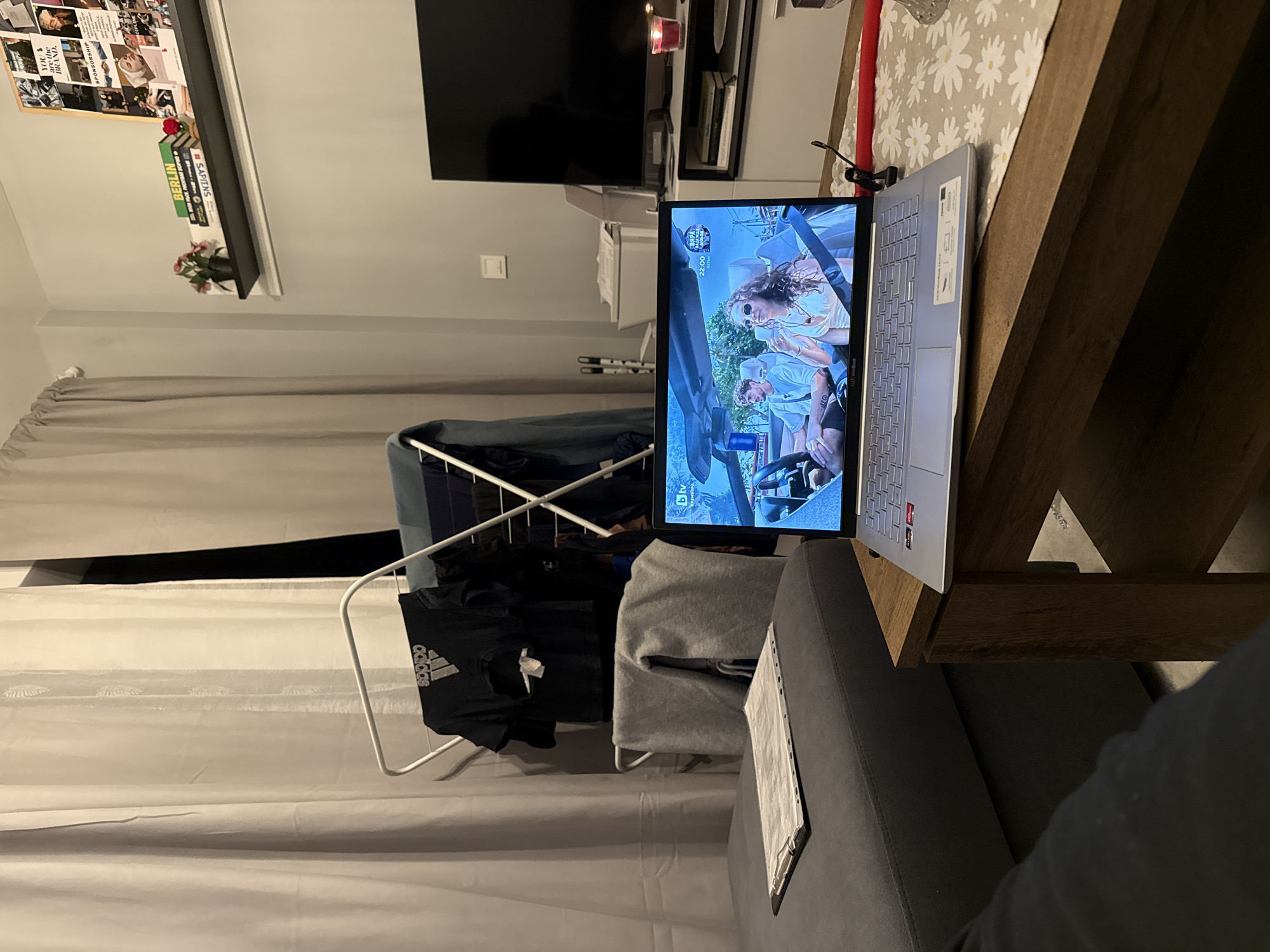 Here is a detailed description of the image:
The image is taken indoors in what appears to be a living room, possibly located in Sofia, Bulgaria. The scene is dominated by the presence of a laptop positioned on a wooden table next to a gray couch. On the laptop screen, a program is playing; the details suggest that it may be a show or a movie from a Bulgarian TV channel named BTV. Based on the on-screen text, the program includes a show called "ВЯРА", playing at 22:00.
In the background, a drying rack holds several pieces of clothing, including a black Adidas t-shirt and a gray garment. The curtains are drawn, and a television set is mounted on the wall, above a cabinet. On the wall shelf, there are several books including the book "BERLIN" and "SAPIENS", along with a small decorative element. There is also an office or personal board, as well as a vase with flowers.
The lighting seems to be mixed with artificial light. The scene has a casual and lived-in feeling, as though someone is spending time at home watching TV or working on their computer. A part of a person's arm, dressed in a dark sleeve, is seen on the bottom right side.