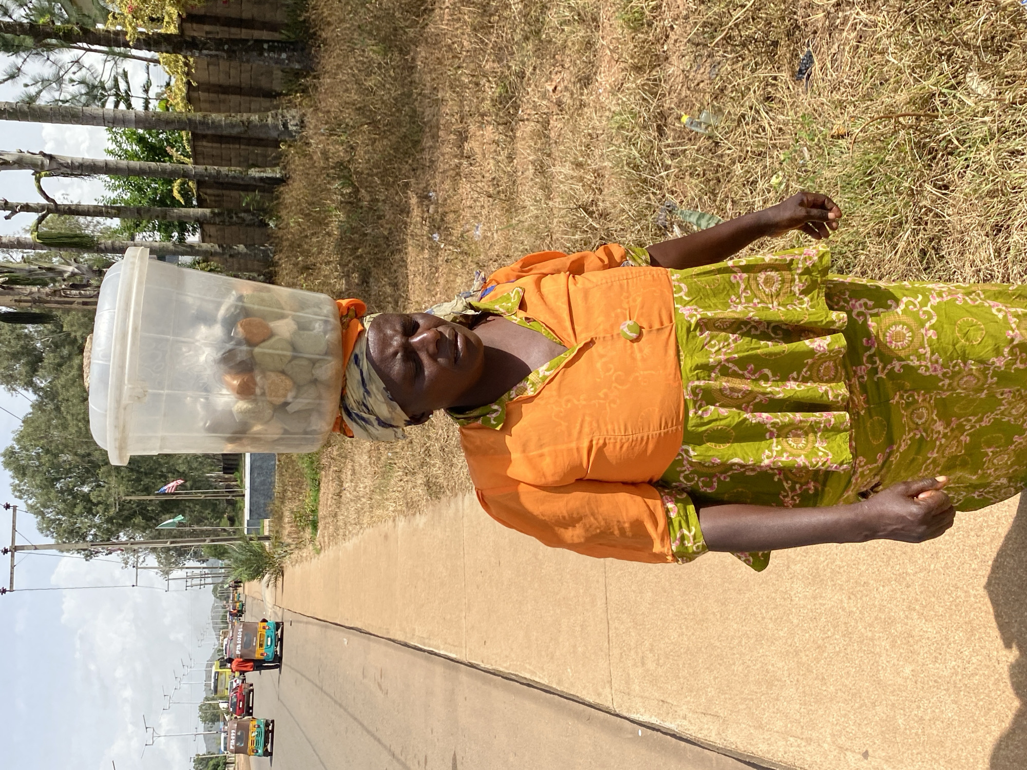 This media file shows a woman carrying a clear plastic container filled with various items on her head. She is wearing an orange top over a green patterned dress and a headscarf. The woman appears to be standing on a paved surface next to a dry, grassy area. In the background, there are palm trees, a wall, and a road with several auto-rickshaws traveling in the same direction. Utility poles with wires are also visible along the road, and flags can be seen in the distance. The scene is brightly lit, suggesting it is daytime, and the weather appears to be sunny. The auto-rickshaws have numbers on them, such as "PB: 1040S" and "PB: 10099". The overall setting appears to be a roadside in a developing area, likely Bukuru, Nigeria. The woman's expression is neutral.