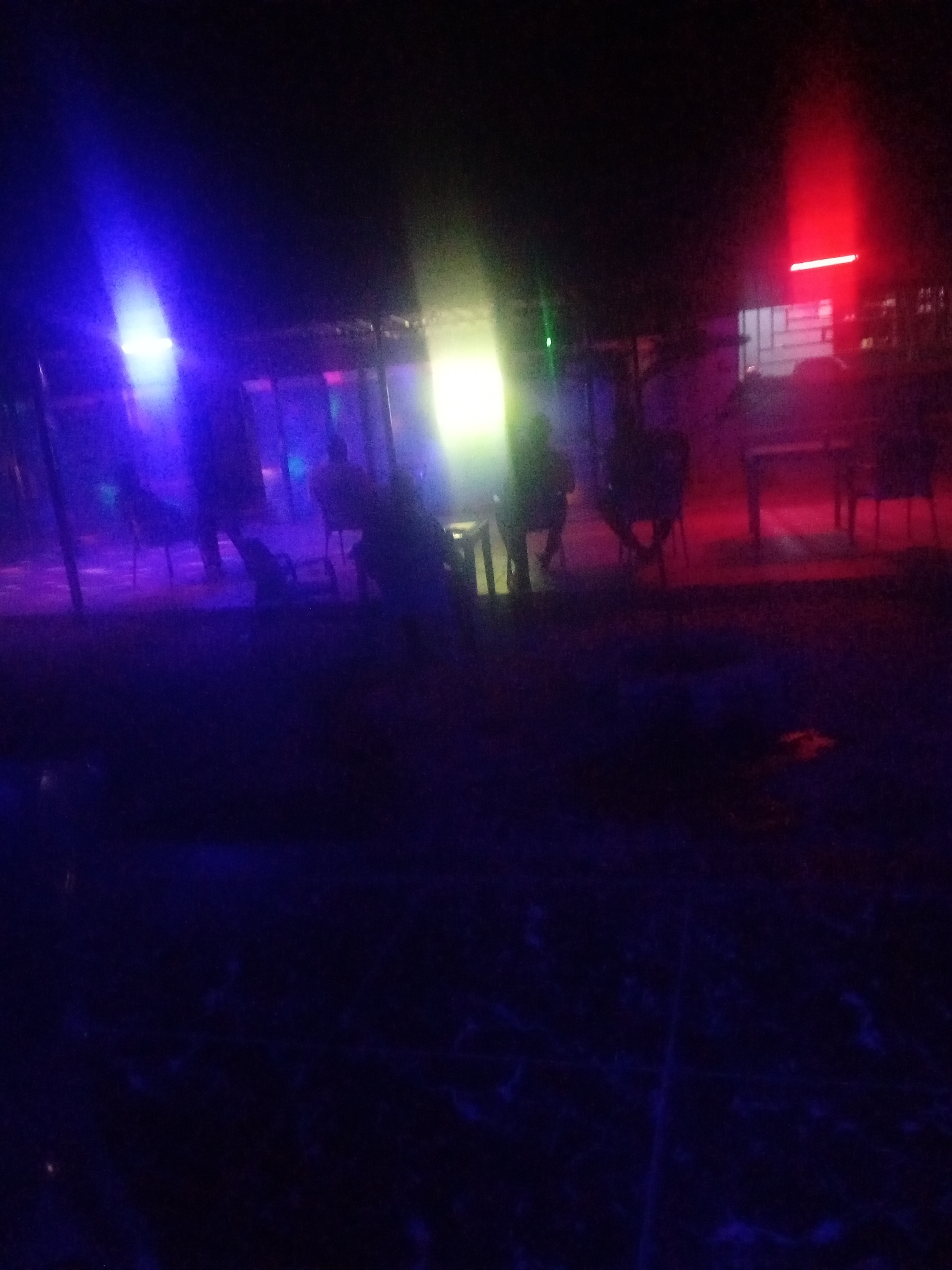 An outdoor nighttime scene at an unspecified location in the city of Unknown, Unknown, is depicted with vibrant colored lighting and silhouetted figures. In the background, a bright red light emanates from a structure with horizontal and vertical bars, possibly a window or gate. To the left, a bright blue light illuminates a portion of the scene, and a bright yellow-green light is visible in the center. Several figures are present, appearing as dark silhouettes against the colored lights. Some figures are seated, suggesting they are at tables or on chairs. A structure with a roof and poles is visible, implying an outdoor seating area or canopy. The ground appears to be a dark, possibly paved surface, with some unevenness. The overall atmosphere is that of a dimly lit outdoor gathering or establishment, with the colored lights creating a dramatic and somewhat abstract effect.