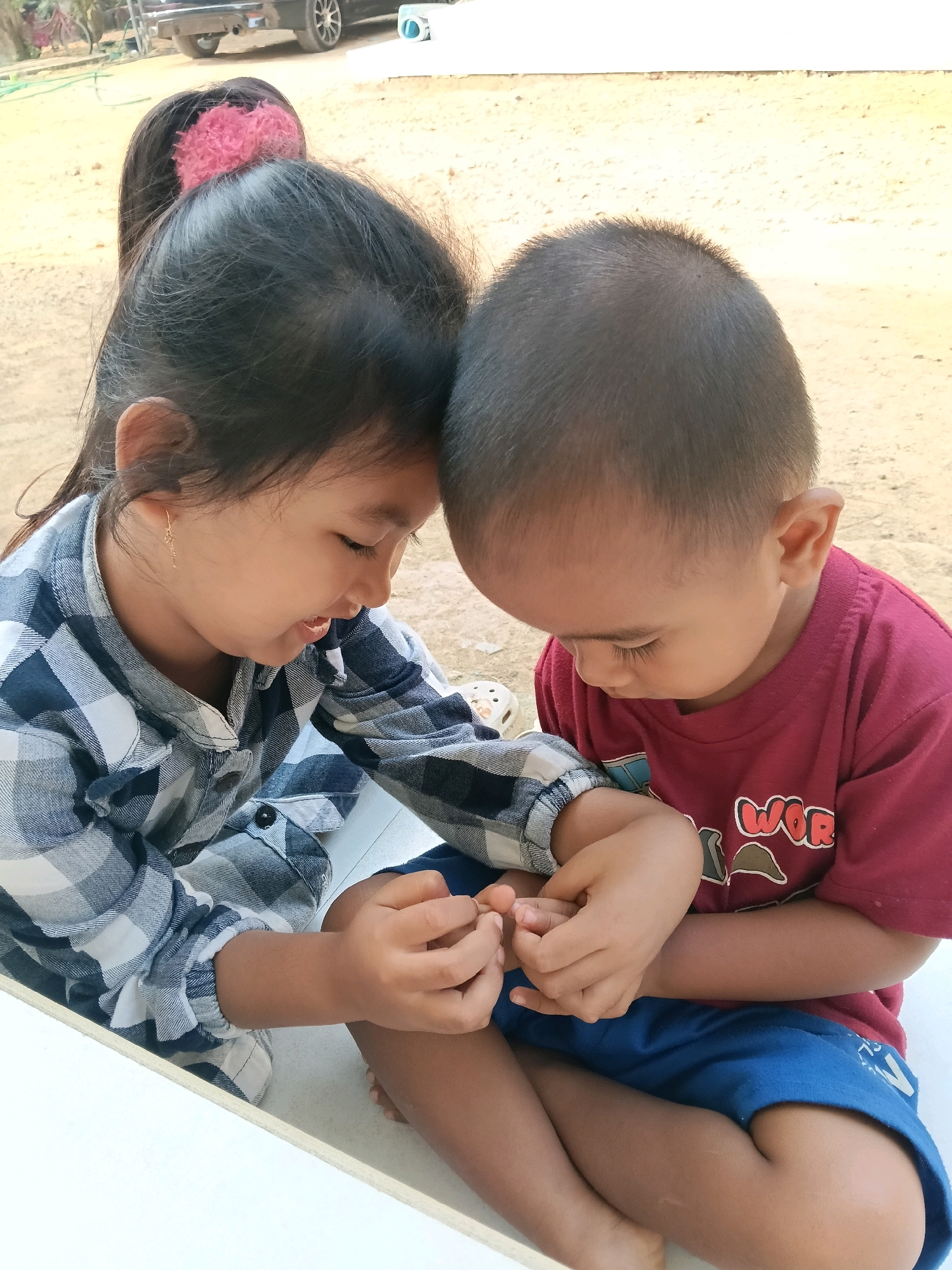 Two young children, a girl and a boy, are seated close together with their foreheads touching. The girl, on the left, wears a blue and white checkered shirt and has her dark hair pulled back with a pink scrunchie. She is looking down and smiling. The boy, on the right, is wearing a maroon t-shirt with "WORLD" printed on it in red letters and blue shorts. He is also looking down, his expression unreadable. They are holding hands, their fingers intertwined. The setting appears to be outdoors, with a sandy or dirt ground visible in the background. The lighting suggests it is daytime.