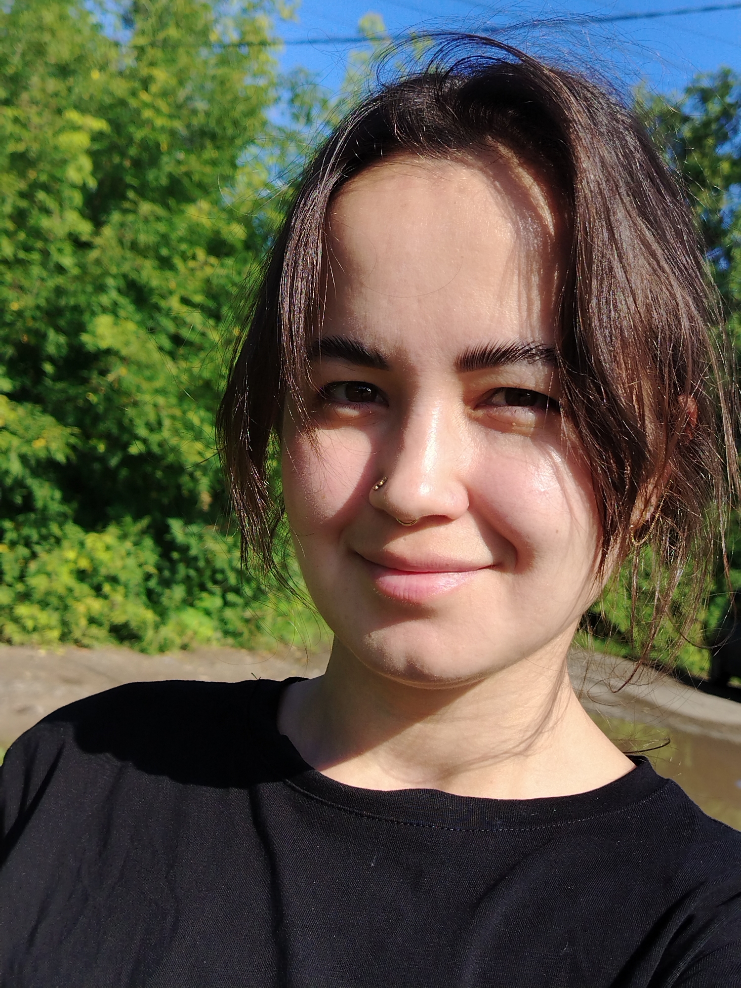A medium shot captures a young woman in a black t-shirt, looking towards the camera with a slight smile. She has dark, shoulder-length hair with bangs, dark eyebrows, and a nose ring. The background features blurred green foliage under a bright blue sky. The lighting suggests it is daytime, with sunlight casting shadows on the woman's face and shirt. The environment appears to be outdoors, possibly in a park or natural setting. There is no indication of the specific location within Kuyeda, Russia.