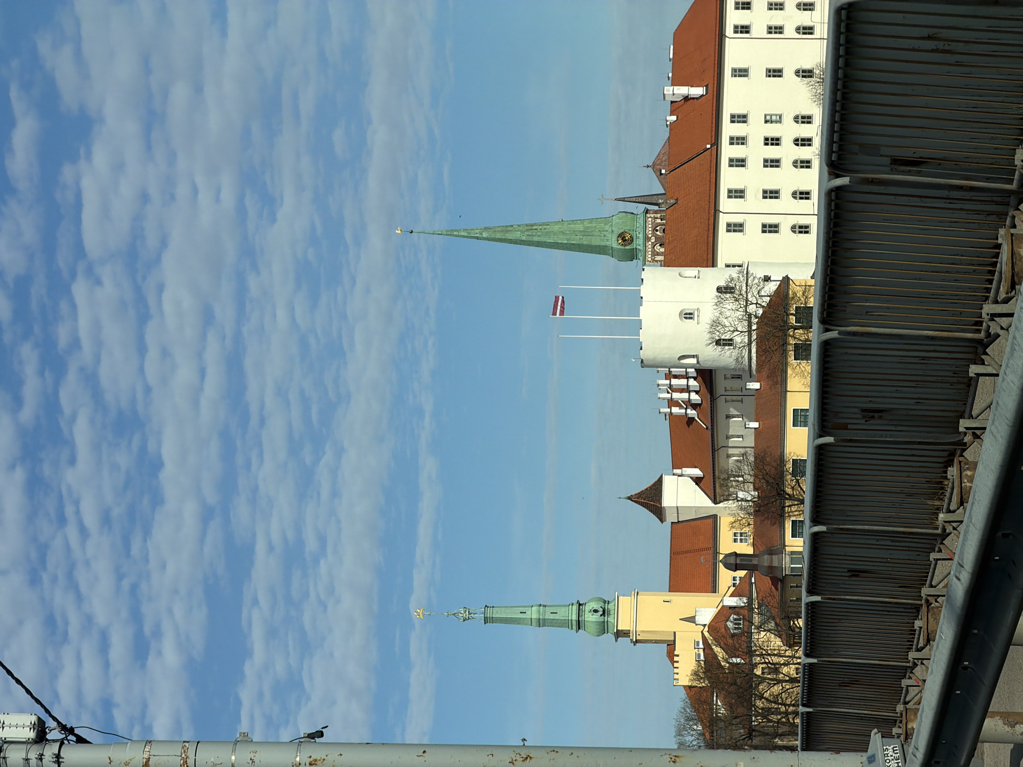 A daytime view of a cityscape in Riga, Latvia, is captured under a bright blue sky with scattered, elongated white clouds. The scene is dominated by several multi-story historical buildings featuring light-colored facades, including white, cream, and yellow, topped with distinctive red tiled roofs. Many windows are visible, with both arched and rectangular shapes.
Two prominent green spires, likely belonging to churches, rise towards the sky. The more central spire features a clock face and is topped with a golden weathercock. Another green spire with a clock is visible further to the right. Near the central spire and a white tower, two Latvian flags with their characteristic red-white-red stripes are seen flying from flagpoles, indicating the national context. Bare deciduous trees are interspersed among the buildings, suggesting a late winter or early spring season.
In the foreground and along the right edge, a weathered metal barrier or railing, possibly from a bridge or elevated walkway, is prominently featured, showing vertical slats and horizontal bars. This structure partially frames the urban landscape. A small label on this barrier in the bottom right corner displays the text "KORS HEM." The bright lighting suggests a sunny day.