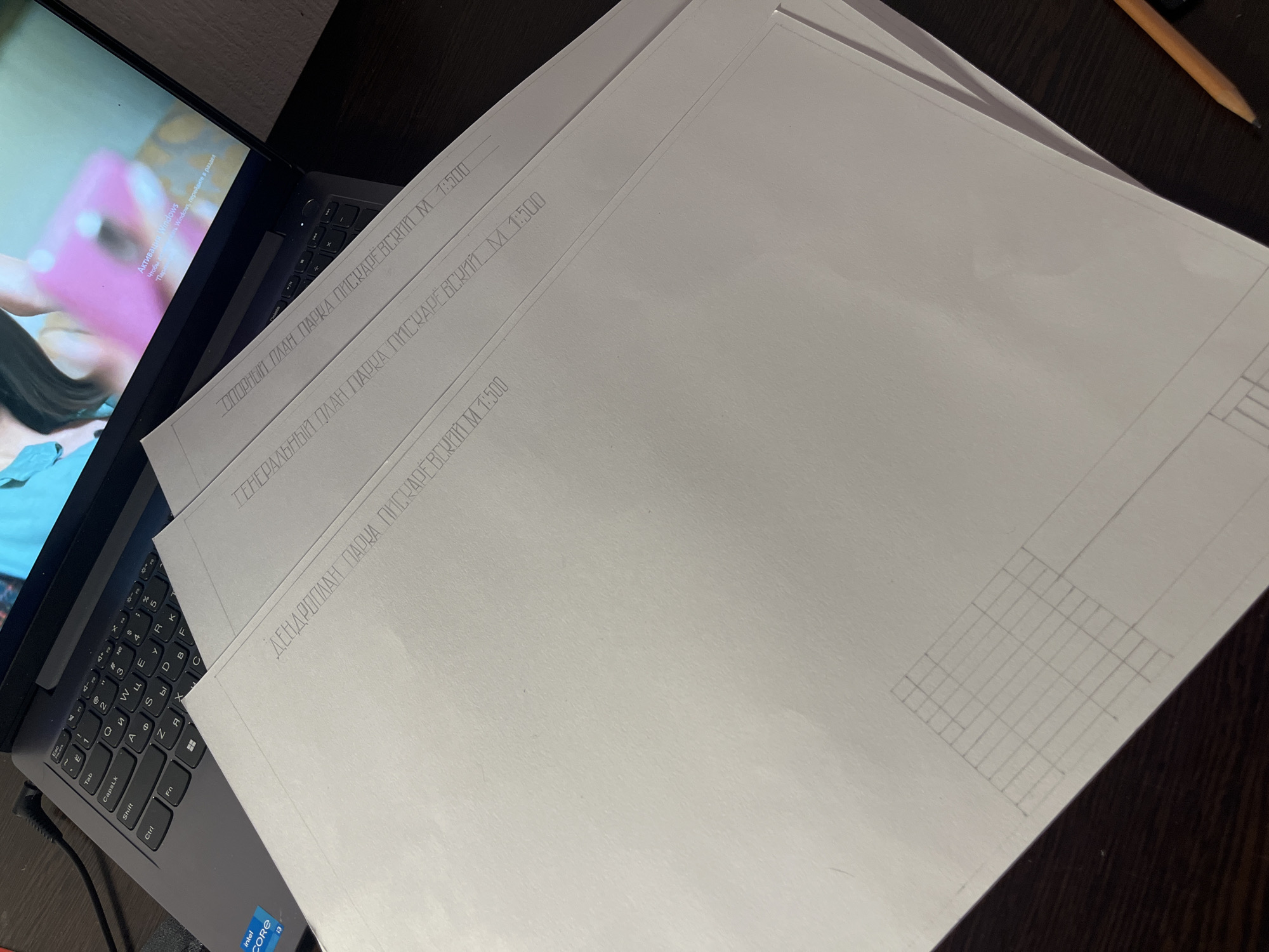 The image shows a cluttered desk with a laptop partially visible in the lower left corner. The screen displays a Windows activation message. Stacked on top of the laptop are three sheets of white paper with technical drawings or architectural plans. The text "ПЛАН ПАРКА ПИСКАРЕВСКИЙ M 1:500" (which translates to "Piskarevsky Park Plan M 1:500") is printed on the edge of each sheet. The uppermost sheet has a hand-drawn grid visible in the lower right corner. A pencil lies on the desk near the upper right corner. The scene is dimly lit and the background is a dark surface.