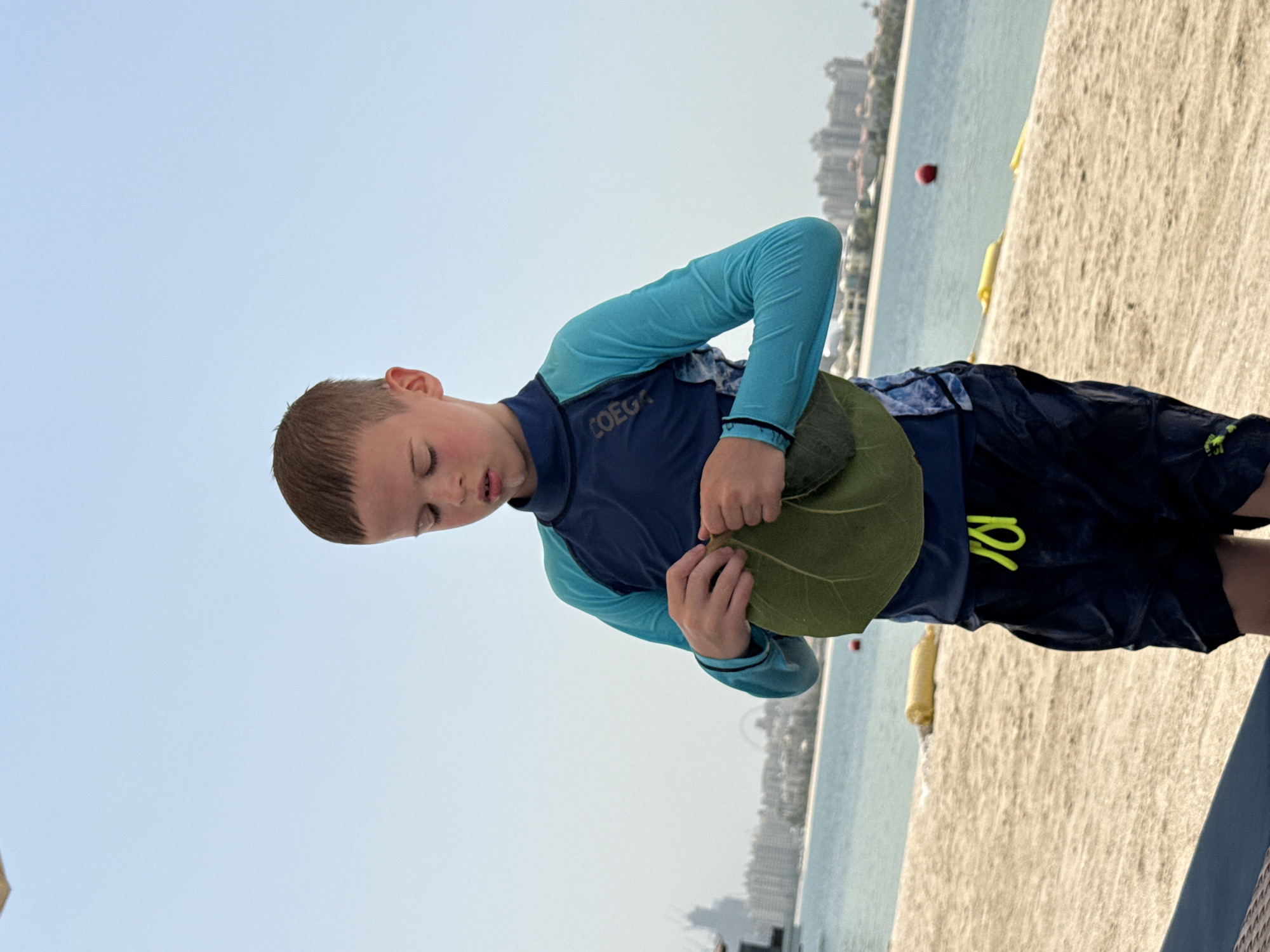 A young boy, wearing a blue and teal rash guard and black swim shorts with a neon yellow drawstring, stands on a sandy beach holding a large green leaf. The boy's eyes are downcast as he examines the leaf, his fingers manipulating it. In the background, the turquoise waters of the Arabian Gulf meet a hazy sky. A distant cityscape of Dubai, characterized by modern architecture and buildings, is visible along the coastline, with a tall tower and Ferris wheel suggesting the presence of JBR or Palm Jumeirah. Yellow buoys float in the water, and a red buoy is visible further out. The beach appears to be part of a resort or public area, with some beach furniture partially visible in the foreground. The overall scene captures a moment of quiet observation on a bright, warm day.