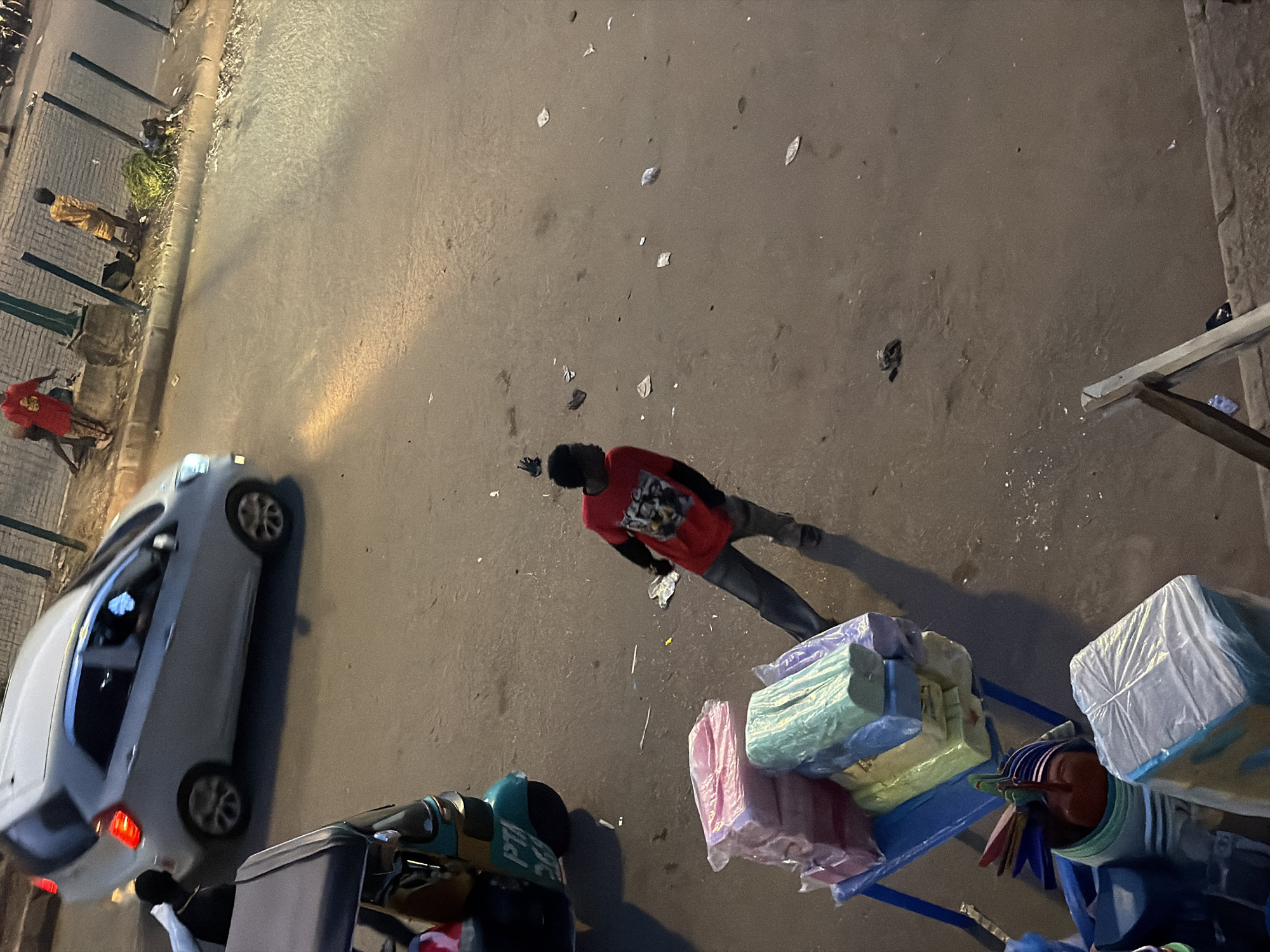 A street scene in Bukuru, Nigeria, depicts a dusty, paved road with a slight incline. A white sedan is in motion on the left, its brake lights illuminated. A young man in a red t-shirt with a graphic print and dark pants stands in the middle of the road, facing away from the camera. To his left, a turquoise and yellow auto-rickshaw is parked. In the foreground, a blue table holds stacks of colorful items wrapped in plastic, likely merchandise. The ground is littered with debris, including paper and plastic waste. In the background, a concrete sidewalk and a fence line are visible, with two individuals standing near the fence. The lighting suggests it is either early morning or late evening.
