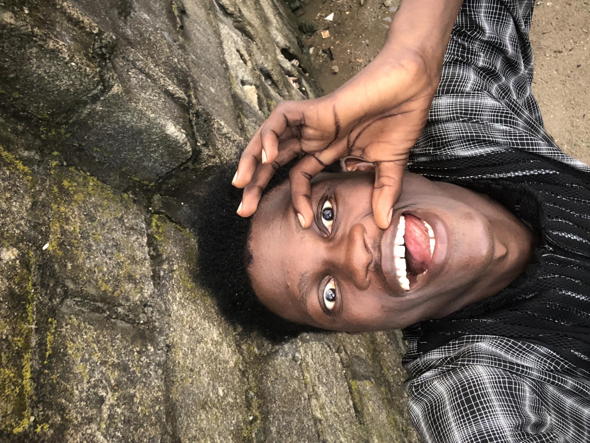 An overhead shot captures a young Black man from the chest up, lying down outdoors. He is looking directly at the camera with a playful expression, his mouth wide open and tongue slightly visible. His left hand is raised, with fingers pinching his left eyelid, while his right hand rests on his chin. He has dark, curly hair and is wearing a black and white checkered shirt with a black mesh underlayer. The background consists of rough, grey stone with patches of green moss, suggesting a natural or perhaps a rugged urban environment. The ground beneath him appears to be sand or dirt. This setting is consistent with the rocky and natural areas found on the outskirts of Ado, Nigeria, where one might find such textures and terrain.