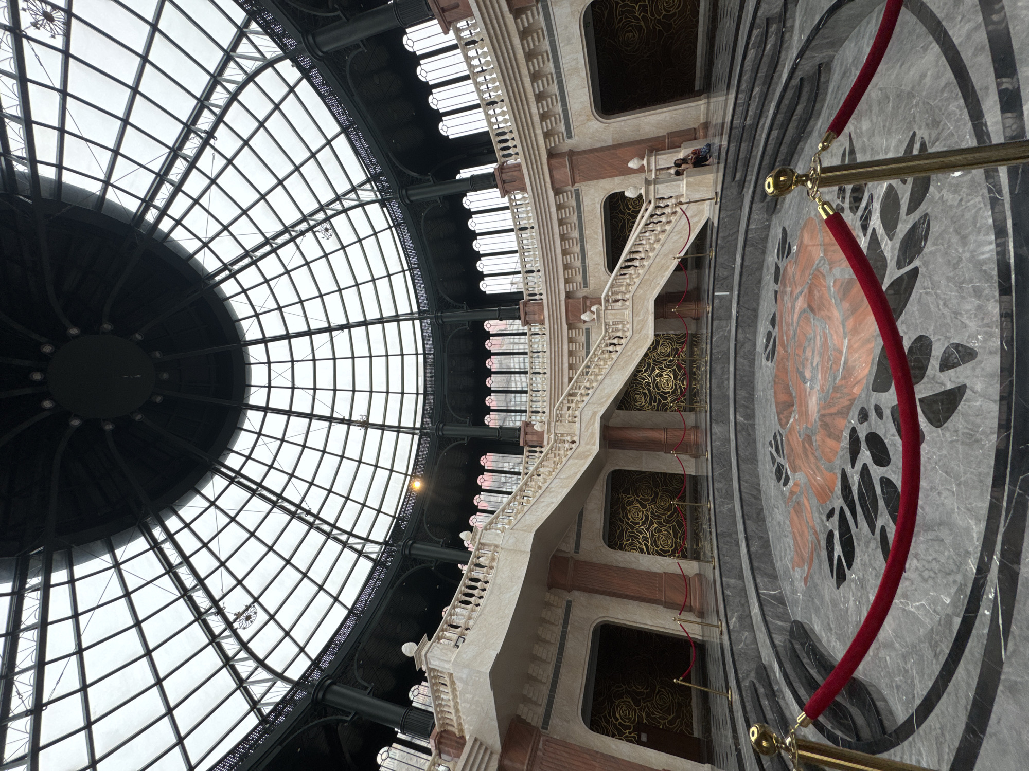 The image captures the interior of a grand building in Đà Nẵng, Vietnam, characterized by elaborate architectural details. A large, domed glass ceiling with a grid-like pattern dominates the upper portion of the image. Below the ceiling, a multi-story structure features a marble staircase with ornate railings. The walls are decorated with a patterned design, and there are several windows. A red velvet rope on gold stanchions is visible in the foreground, potentially indicating a restricted area. A decorative rose design is inlaid into the marble floor. Two people are partially visible on the second floor.