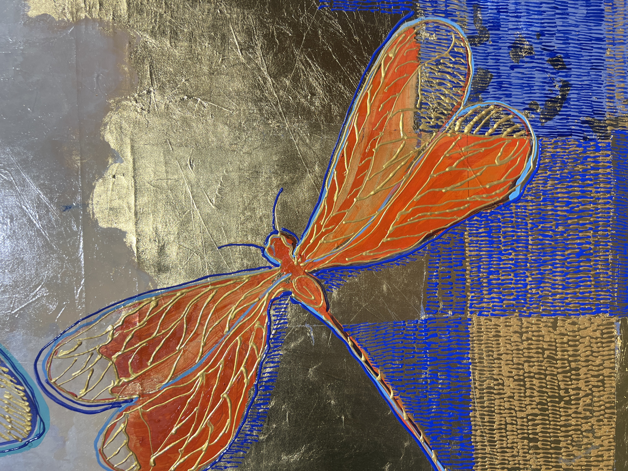 This is a close-up, overhead shot of an abstract painting featuring a stylized dragonfly. The dragonfly, rendered in vibrant orange with gold and blue accents, is the central subject. Its wings are detailed with a network of fine gold lines, suggesting veins. The outline of the dragonfly is in a bold blue, with some sections highlighted in lighter blue.
The background is a textured mix of metallic gold and deep blue. The gold areas appear to have a brushed or hammered metal effect, with varying degrees of sheen and texture, suggesting a rough, reflective surface. Interspersed with the gold are blocks of blue with a raised, ridged texture, creating a checkerboard-like pattern. Some areas of the blue are darker, with hints of black or very dark blue showing through the texture.
The lighting in the image suggests a bright light source, creating strong highlights and shadows on the metallic gold surfaces, which adds to the dimensionality of the artwork. There are no discernible people, specific locations, or a clear narrative of an event taking place. The overall impression is one of artistic expression, with a focus on color, texture, and form. There is no visible text in the image.