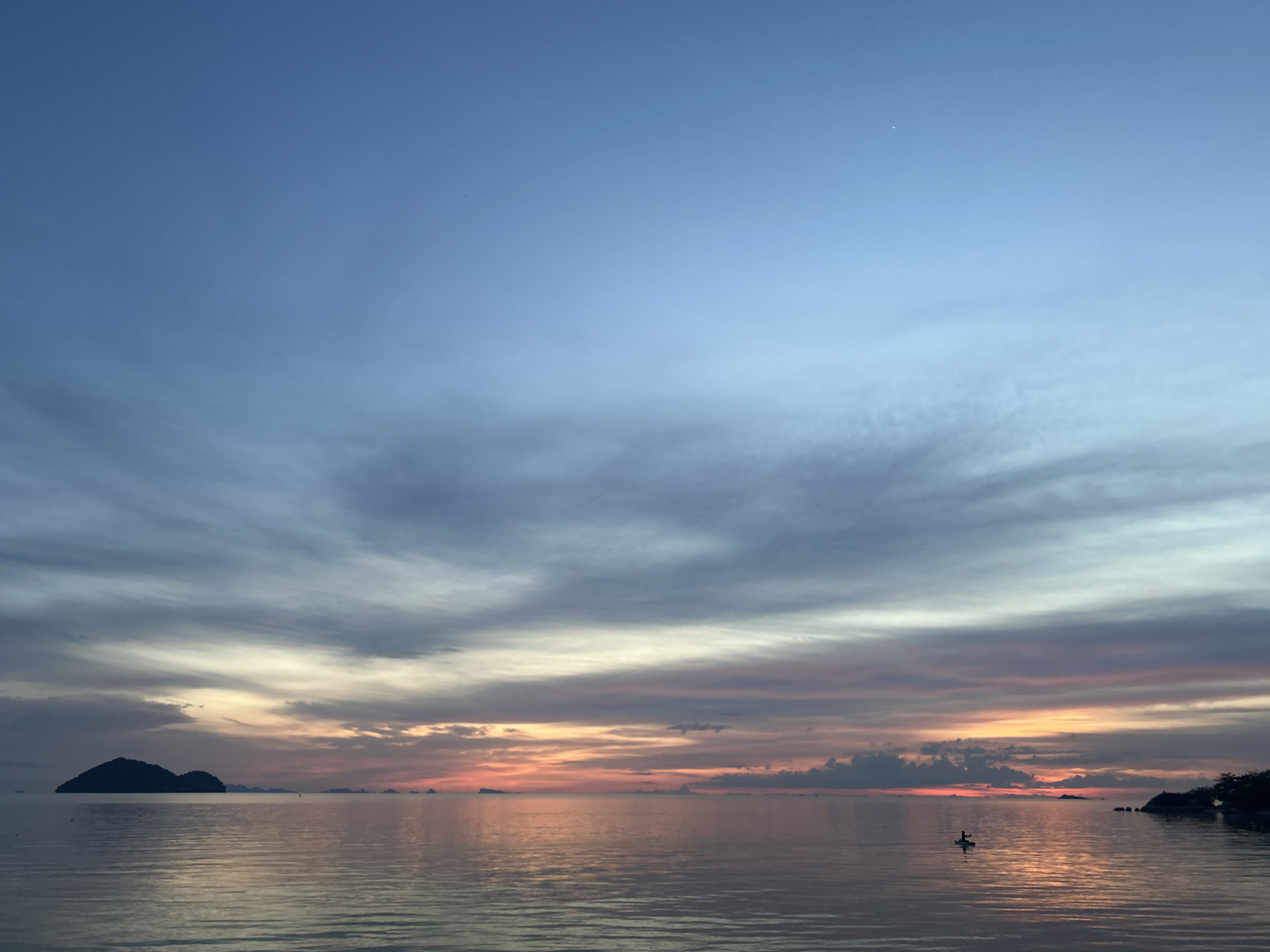 The image depicts a serene sunset over the ocean in Koh Phangan, Thailand. The sky is a gradient of blue, transitioning to hues of pink, orange, and yellow near the horizon, with wispy clouds scattered throughout. The calm sea reflects the colorful sky, creating a mirrored effect. In the distance, several islands are visible as dark silhouettes against the vibrant sky. In the foreground, a lone individual is seen paddling on a board in the water, adding a sense of tranquility and adventure to the scene. The overall atmosphere is peaceful and picturesque.