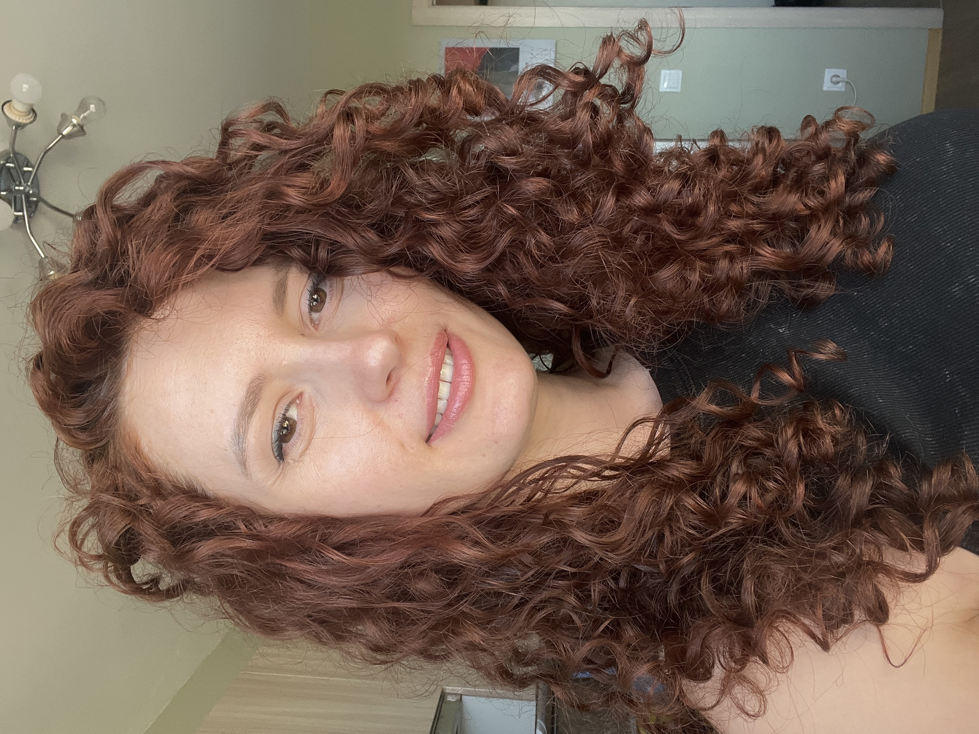 A woman with long, curly auburn hair is facing the camera. She is wearing a black top. The background is a muted green wall with a doorway visible. A light fixture with exposed bulbs is partially visible on the left. The scene appears to be indoors. It is not possible to determine the location of Saint Petersburg, Russia, from this image alone.