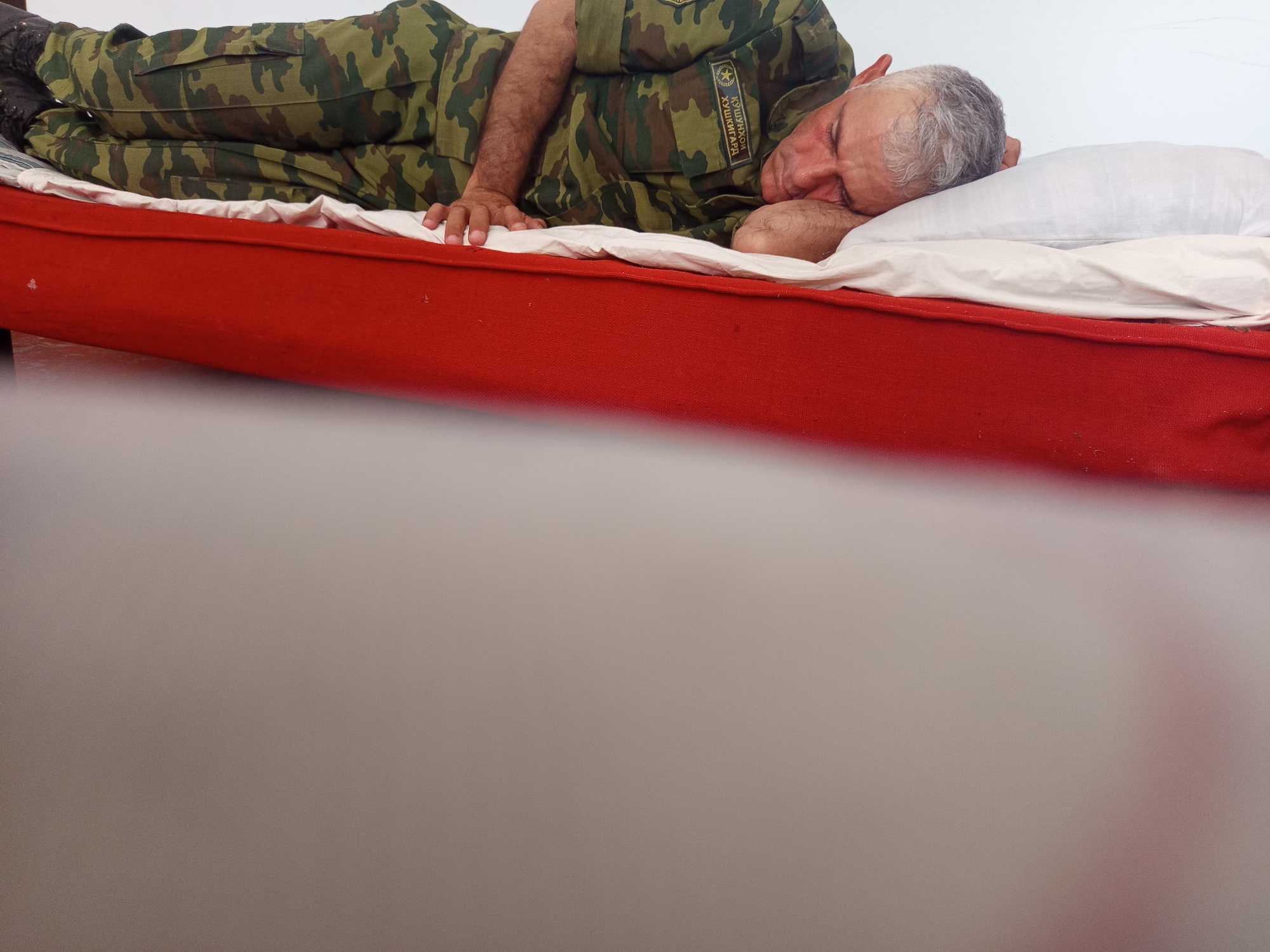 An adult male wearing a camouflage uniform is sleeping on his side on a bed with a red base and white bedding. His head rests on a white pillow, and his eyes are closed. A patch with Cyrillic text and a star emblem is visible on his uniform. The background is a plain white wall. The scene appears to be indoors. No specific location in Buston, Tajikistan is depicted.