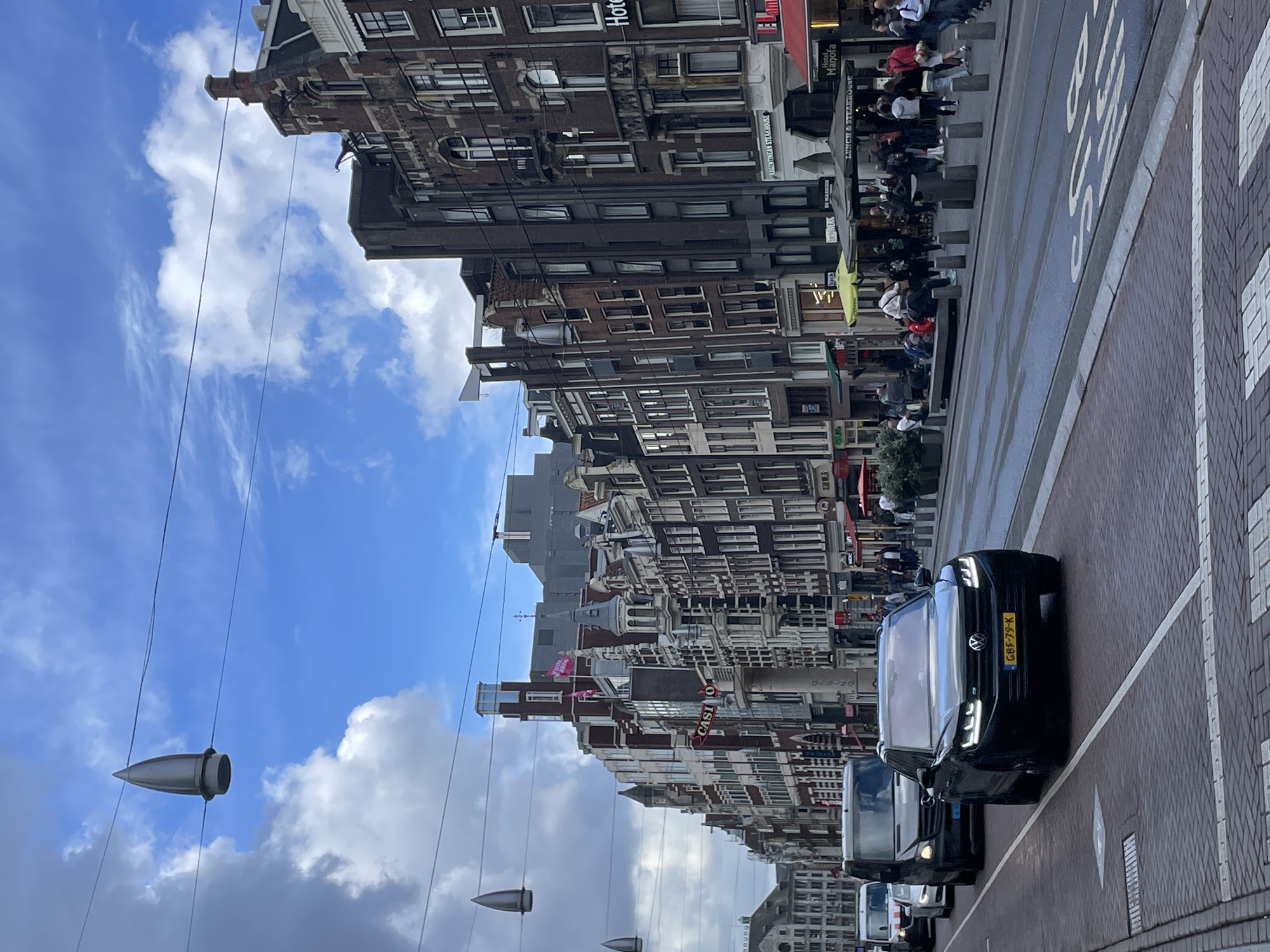 This image captures a vibrant street scene in Amsterdam, Netherlands, on a partly cloudy day. The perspective is looking down a bustling street lined with tall, narrow, historic Dutch buildings.
The main subjects are the distinctive architecture, numerous pedestrians, and vehicles. The buildings feature classic brick facades, gabled roofs, and multiple windows, housing various businesses. Many have awnings and outdoor seating areas, indicating cafes or restaurants. People are visible walking along the sidewalks, dining outdoors, and congregating in groups, suggesting an active urban environment. Several cars are on the road; a prominent black Volkswagen SUV with the Dutch license plate "GBF-79-K" is clearly visible in the foreground, followed by other vehicles.
Overhead, a blue sky with scattered white clouds is visible, crisscrossed by utility wires from which unique, bullet-shaped metallic objects hang, possibly part of a tram system or decorative street lighting. The street itself is paved with a mix of asphalt and brick, featuring clear white road markings, including partially visible text "BUS S".
Visible text on the buildings includes "ARGENTINEAN STEAKHOUSE," "Hotel Manofa," and "LUNCH & STEAKHOUSE." Other partial texts like "Hotel," "CASI" (likely "CASINO"), and "NBURG" (possibly part of a longer word or business name) are also discernible. The overall activity depicts a typical day in a lively city center, with a mix of commercial and pedestrian traffic.