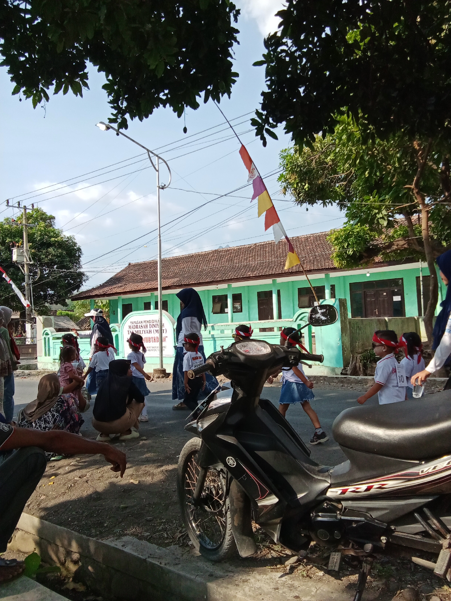 In Trebungan, Indonesia, a group of young children in red headbands are participating in an outdoor activity in front of a turquoise building with a brown-tiled roof. The children, dressed in white shirts and some in denim skirts, appear to be a part of a school event, possibly a procession or game. One child wears a shirt with the number "30" visible. Several adults are present, some observing the children and others seeming to guide them. A person in a hijab is visible on the right side of the frame, holding a clear bottle. In the foreground, a black motorcycle is parked, partially obscuring the lower portion of the scene. The environment includes trees with lush green leaves providing shade, a street lamp, power lines, and a street sign indicating "YAYASAN PENDIDIKAN ISLAM MADRASAH DINIYAH TAKMILUYAH (MI) NURUL HUBUTAN," which identifies the institution as an Islamic education foundation. The ground is paved with asphalt and dirt. The sky is clear and blue.