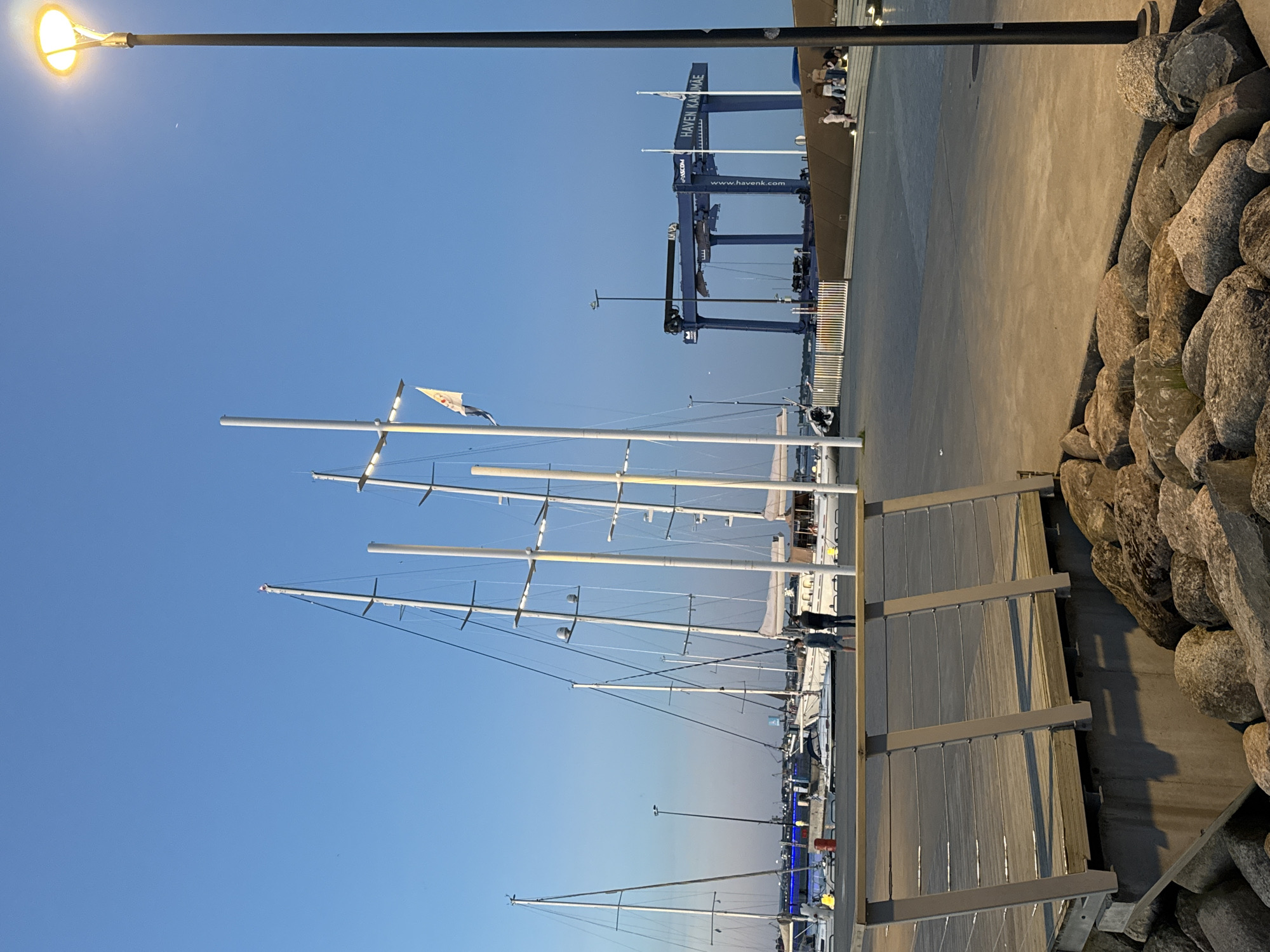 This is an outdoor, eye-level, medium shot of a harbor area in Tallinn, Estonia, at dusk. The sky is a clear, deep blue with a single, faint crescent moon visible. A brightly lit street lamp stands on the right side of the frame, casting a warm glow. In the foreground, a wooden boardwalk with a metal railing leads towards the harbor. The boardwalk is bordered by large, natural rocks.
The middle ground of the image is dominated by the masts of several large sailboats docked in the harbor. The boats are white and appear to be modern yachts. Beyond the sailboats, a large blue boat lift, branded with "ASCOM" and "HAVEN KAKUMÄE", stands prominently. This boat lift is a key indicator of the location. Several people are scattered throughout the scene: two individuals stand on the dock between the boats, and a small group of people are seated on steps to the right of the boat lift. The water in the harbor is calm and reflects the lights from the dock. The overall impression is one of a peaceful evening at a modern marina.