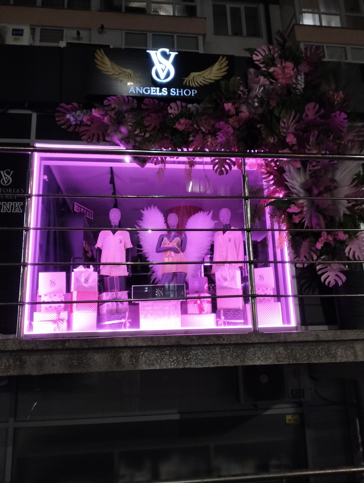 The image shows a shop window of "VS Angels Shop" located in Ivano-Frankivsk, Ukraine. The shop front features a black sign with the logo and name in white letters, adorned with golden wings above. The window display is lit with a vibrant pink light, showcasing mannequins wearing lingerie and angel wings, surrounded by gift boxes and shopping bags bearing the brand logo. The window is decorated with artificial flowers and leaves in shades of pink, white, and green, creating a visually appealing display. The building façade is visible, with windows above the shop front. The photograph appears to be taken at night.