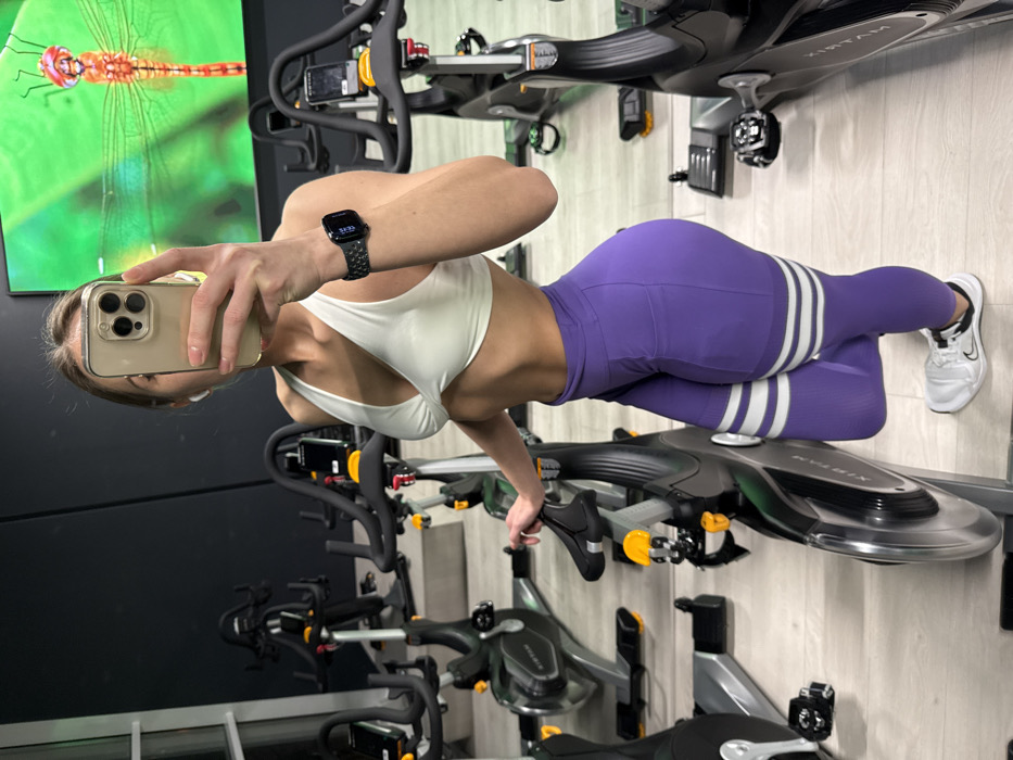 A woman in workout attire poses for a mirror selfie in a gym. She is wearing a white sports bra and purple leggings with white stripes near the ankles. A smartwatch is visible on her wrist. She holds a phone with a tan case in her hand. Behind her are rows of stationary bikes, and a wall with a picture of a dragonfly. The image is slightly rotated.