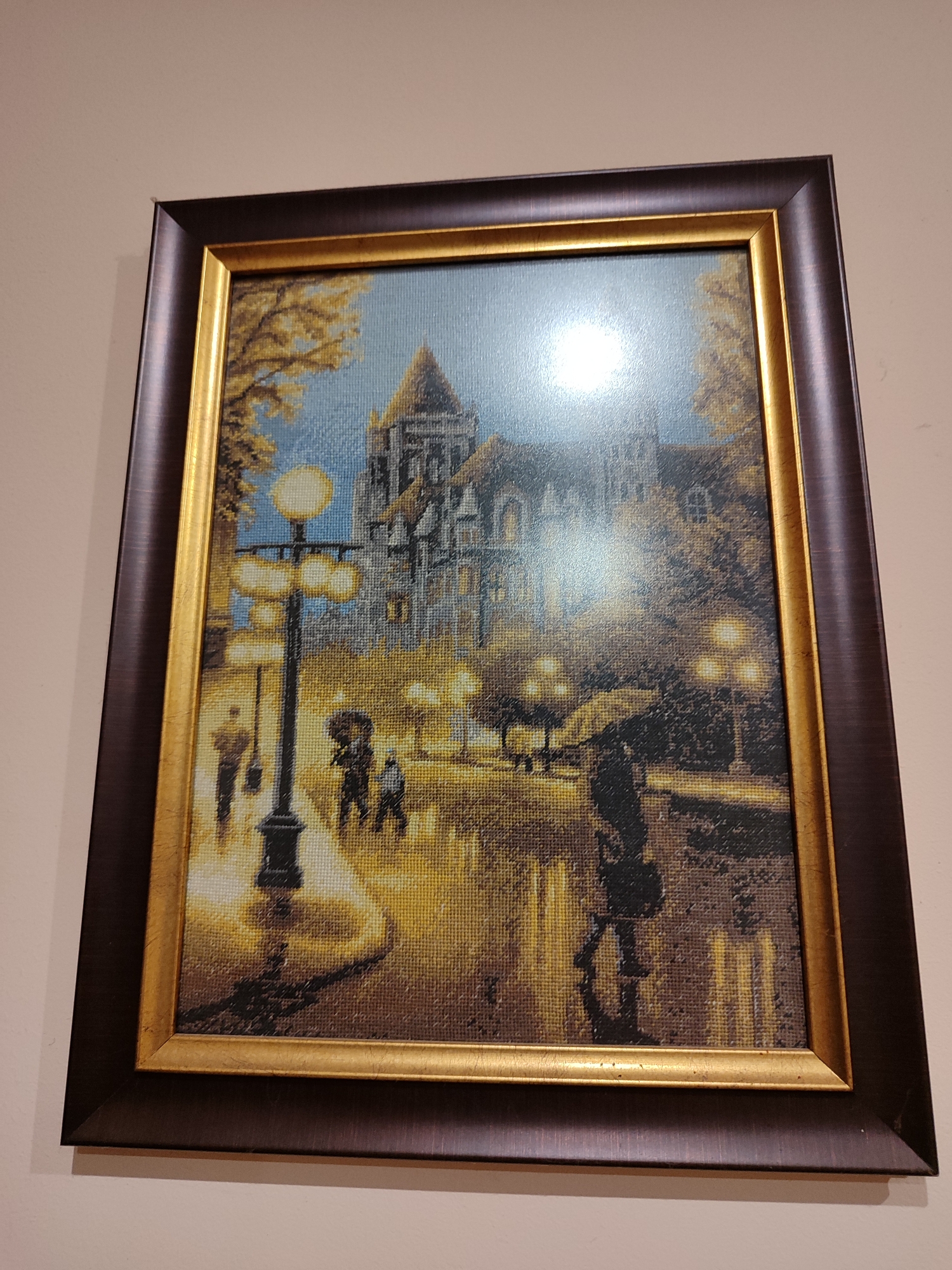 A framed cross-stitch embroidery art depicting a city scene is hung on a light beige wall. The frame is dark brown with a thin gold inner border. The artwork depicts a city street at night or dusk, with people walking with umbrellas, suggesting rain or wet conditions. Street lamps illuminate the scene. A building with a tower is visible in the background. The scene appears to be influenced by an impressionistic style, with a focus on light and atmosphere. It is not possible to determine whether the artwork represents Burgas, Bulgaria.