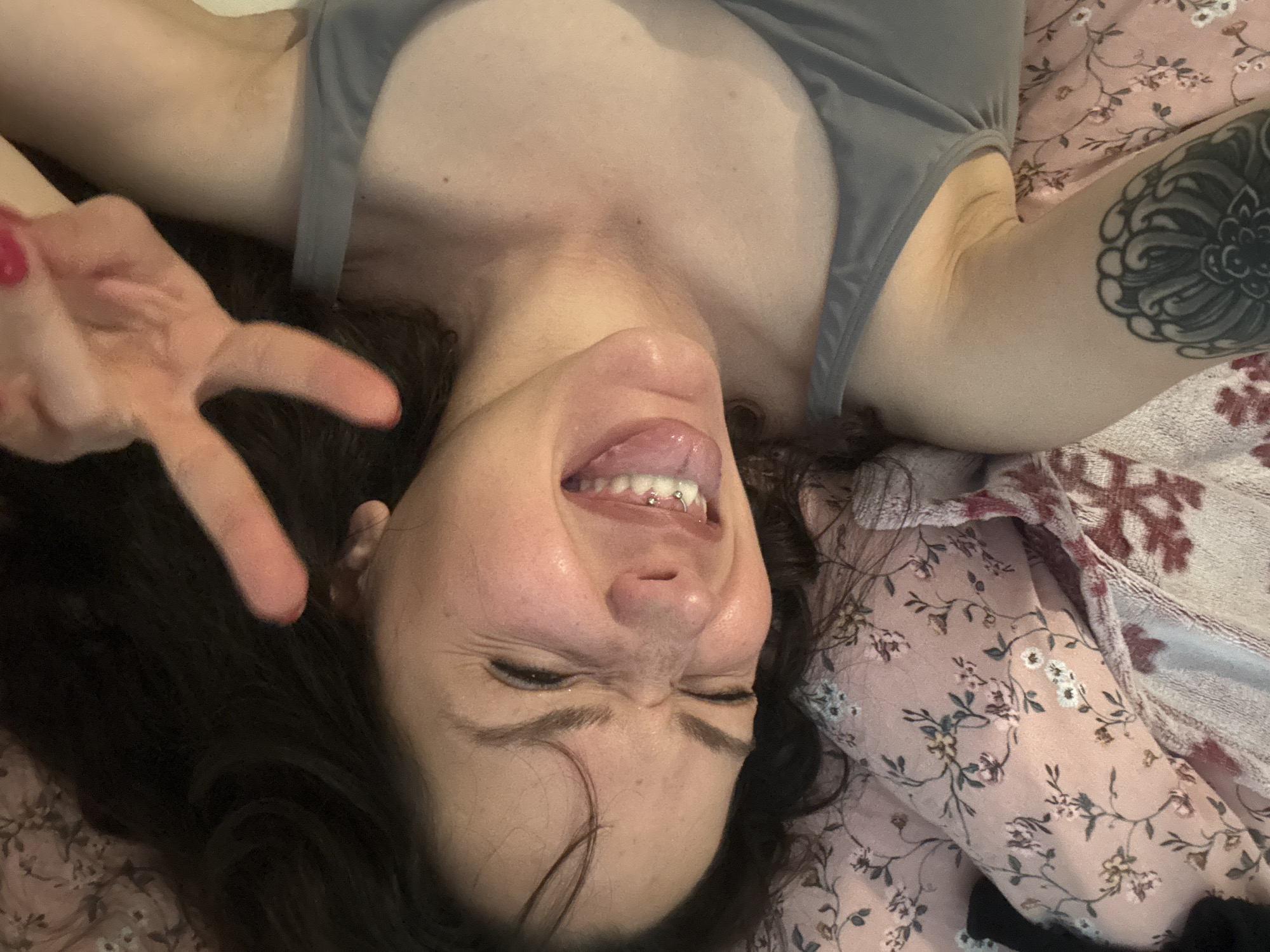 The image shows a person lying on a floral patterned bedspread. The person has dark hair and is wearing a gray tank top. Their right arm is tattooed. The person is making a peace sign with their left hand and sticking their tongue out. The person is smiling with their eyes closed. The tongue has a piercing. There is no indication of the location being in Burgas, Bulgaria.