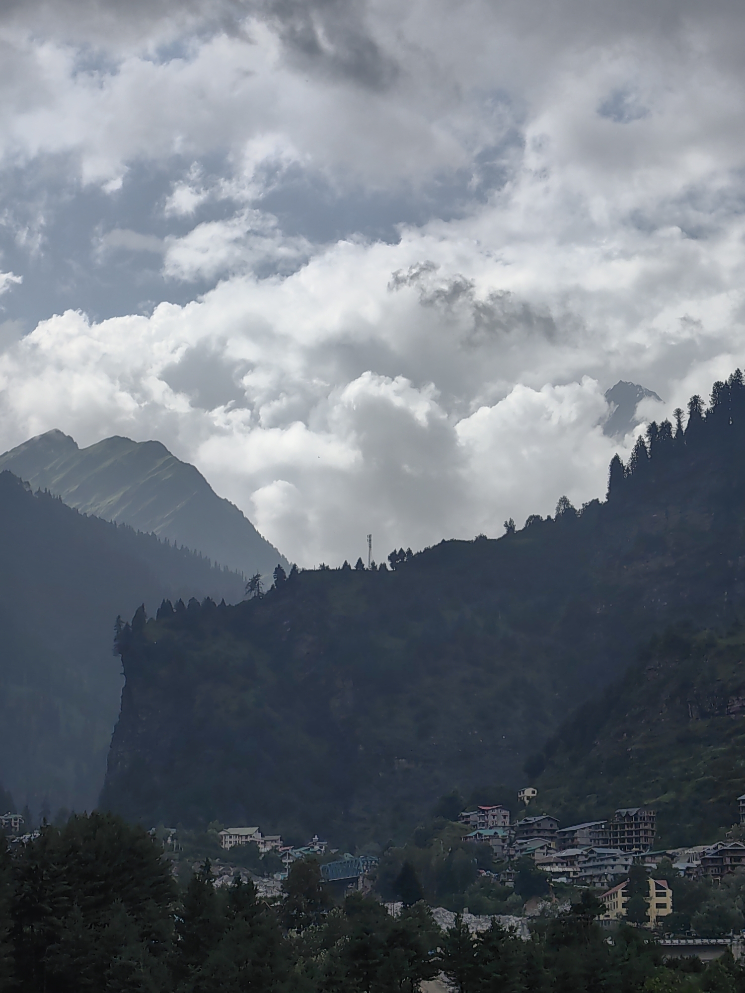 A wide shot captures a cloudy sky over a mountainous landscape and a village in the city of Kullu, India. The sky is filled with dark, dramatic clouds that cast shadows on the mountains. The mountains themselves are steep and covered with dense vegetation. In the foreground, a village is nestled in a valley, with houses and buildings clustered together. A bridge can be seen crossing a river that flows through the village. The overall impression is one of a remote and beautiful natural setting, with a human settlement integrated into the landscape. The image is taken from a high vantage point, looking down on the village. The lighting is dim, suggesting either early morning or late evening, or overcast conditions.